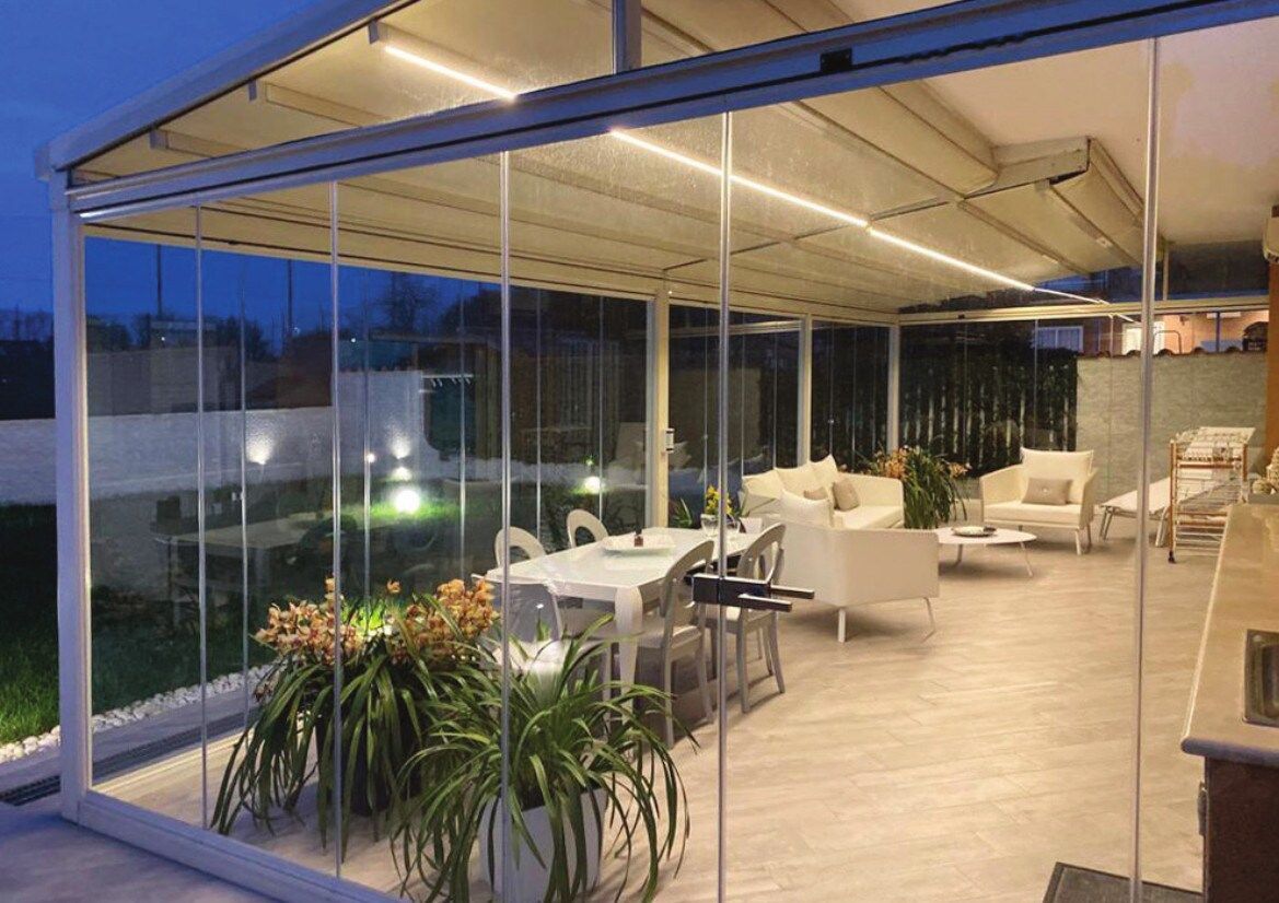 Patio con pareti in vetro, mobili bianchi, tavolo da pranzo, piante e tetto retrattile. Illuminato da luci soffuse.