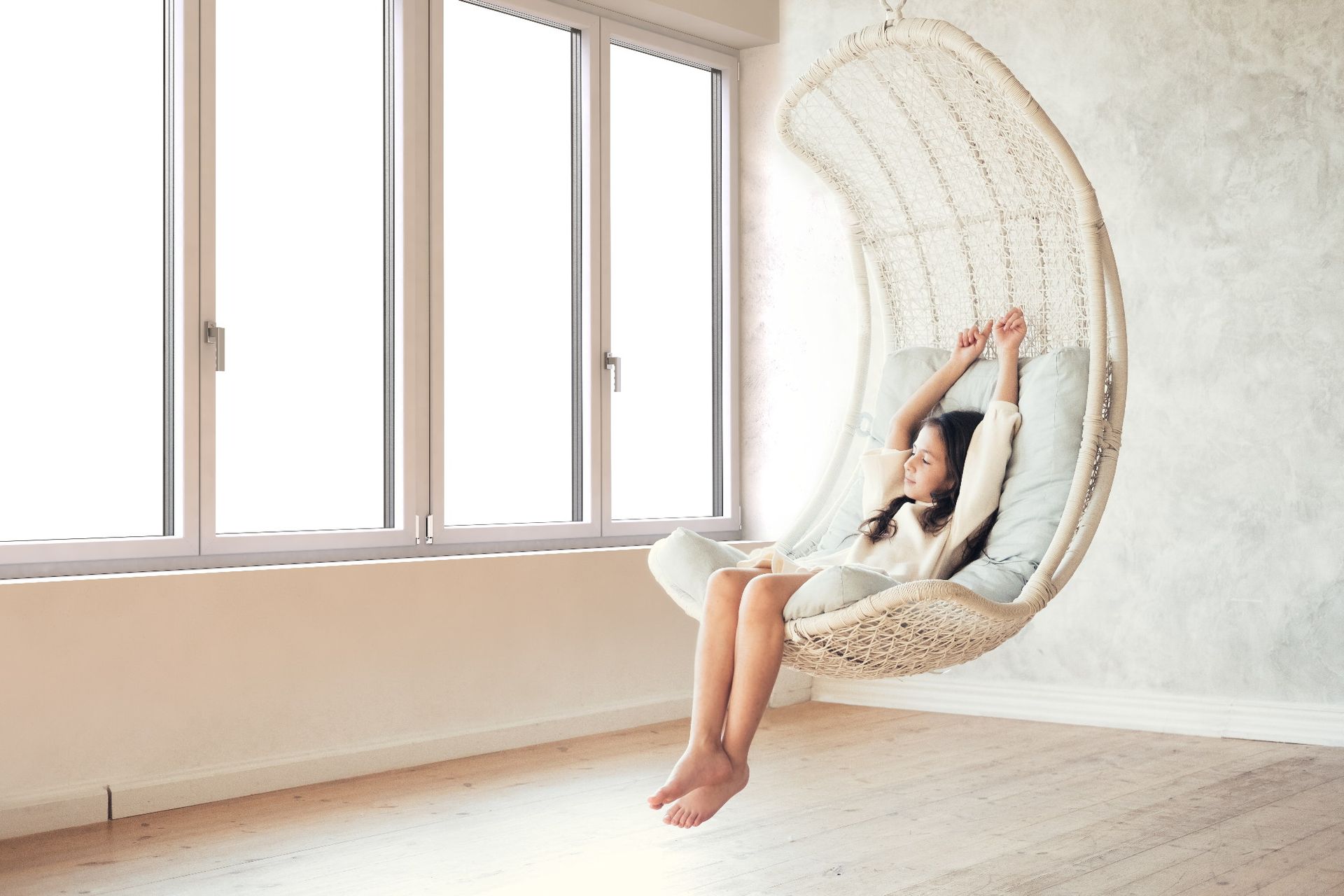 Donna seduta su una sedia sospesa vicino a una finestra, con le braccia alzate, in una stanza luminosa e minimalista.