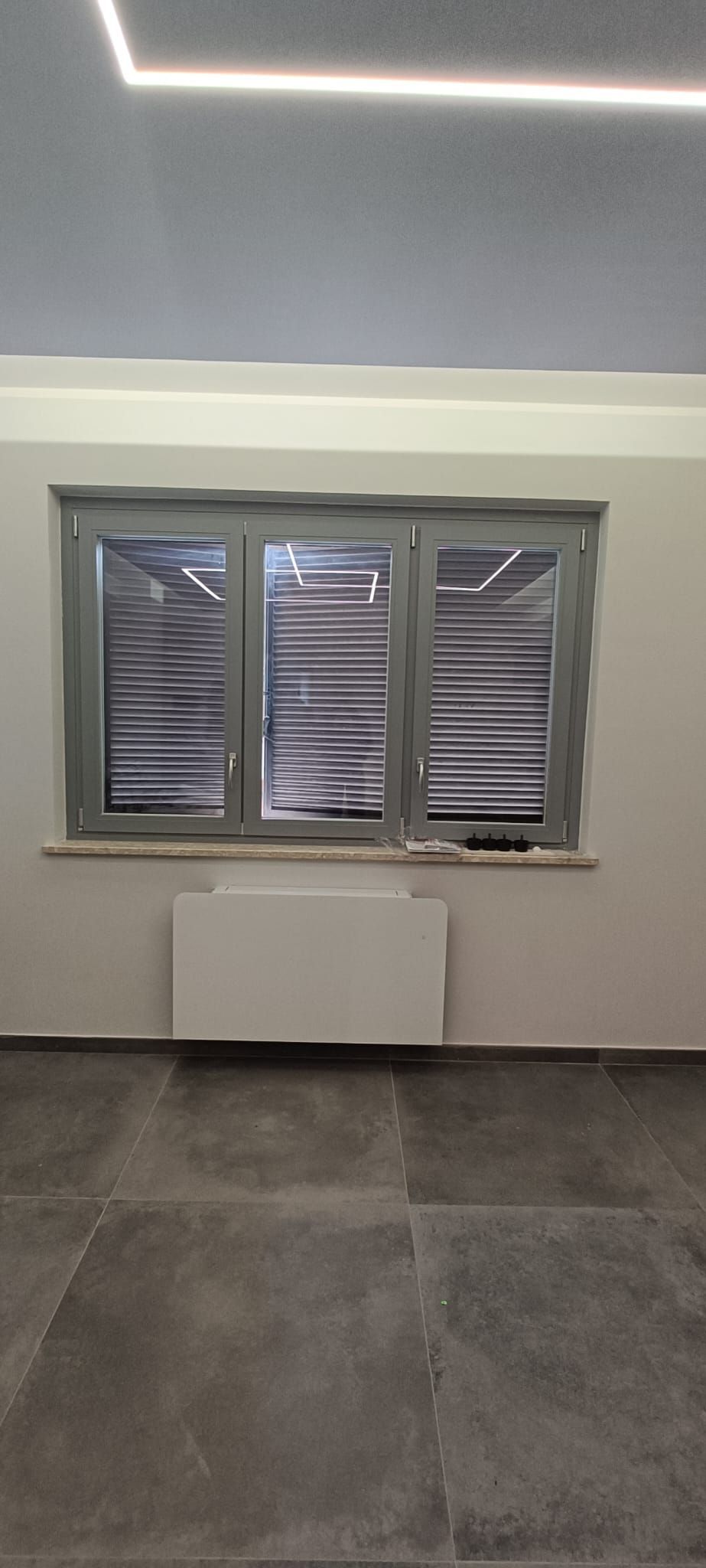 Finestra con persiane chiuse, parete bianca e pavimento piastrellato scuro. Un termosifone bianco rettangolare si trova sotto la finestra.