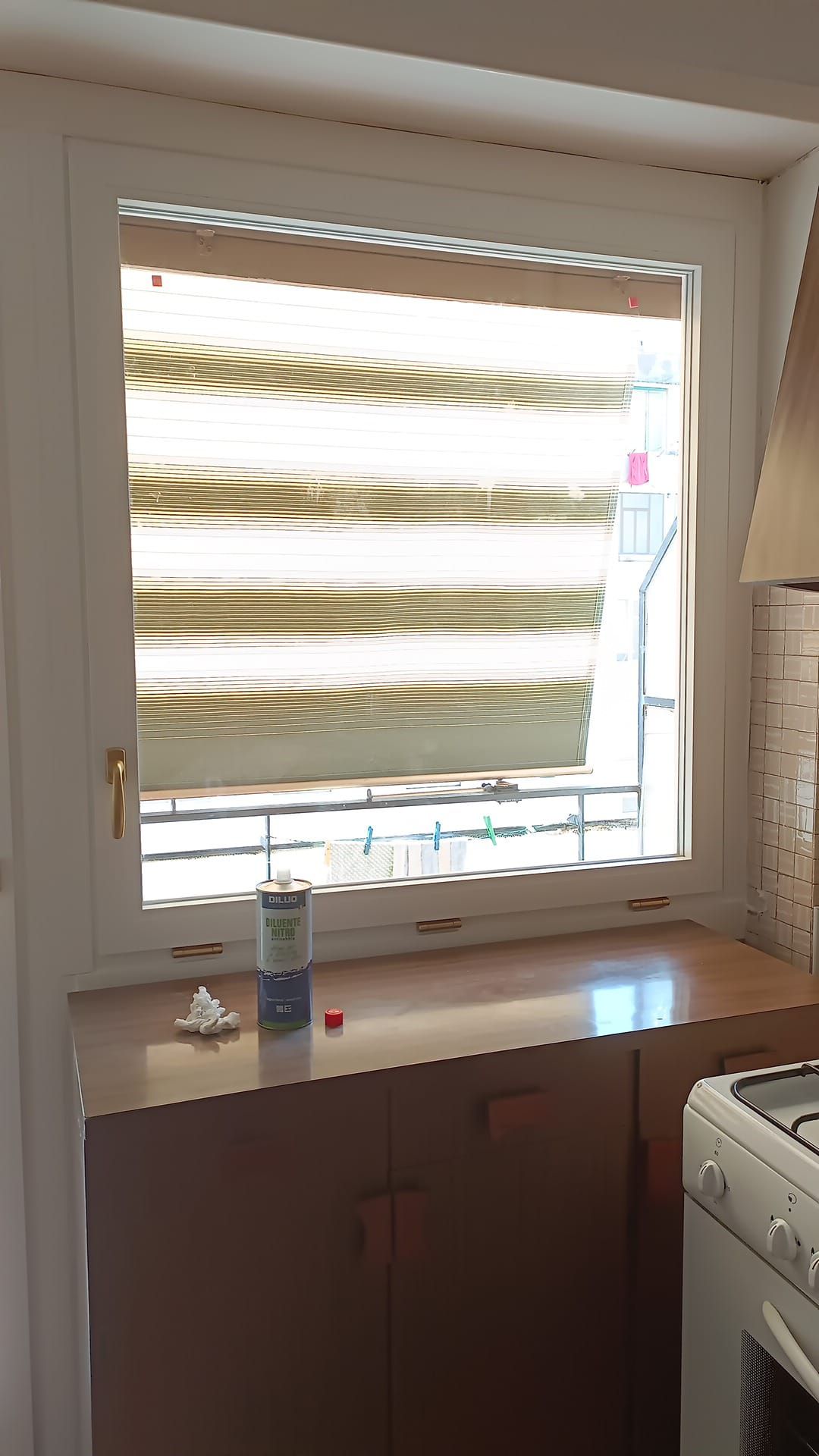 Finestra della cucina con tende a strisce, che si affaccia su un balcone, mobili in basso e fornello a destra.