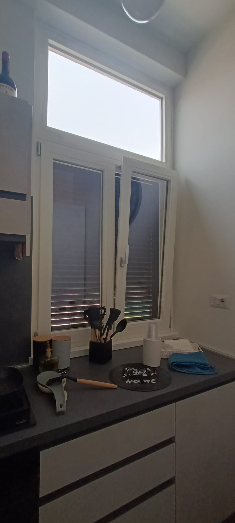 Finestra della cucina con persiane bianche, bancone e oggetti come utensili, tazze e una pianta.