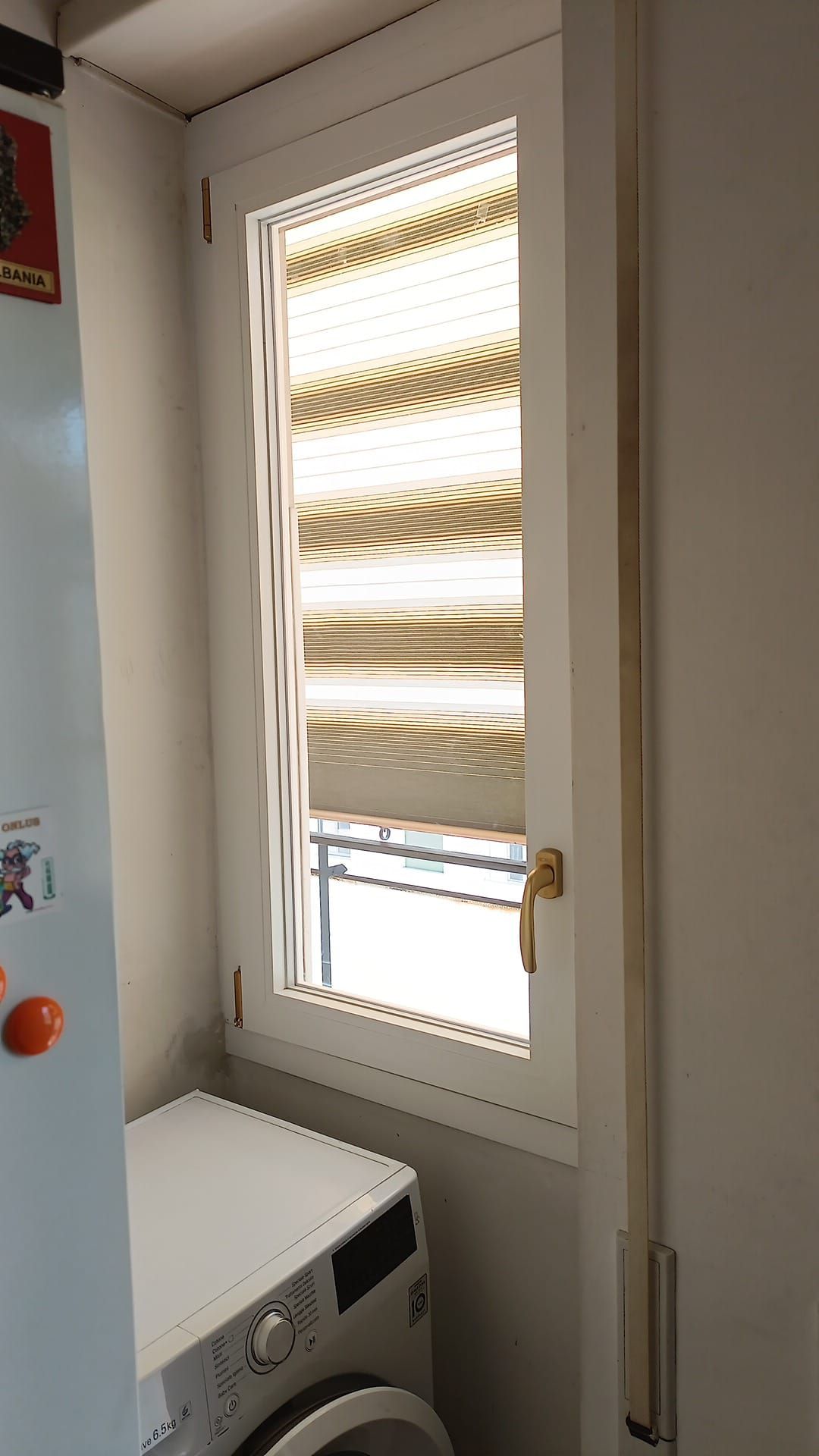 Una finestra stretta con persiane a strisce chiuse sopra una lavatrice in una stanza dalle pareti bianche.