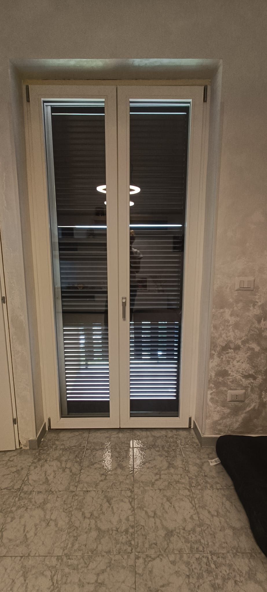 Doppie porte bianche con persiane, incastonate tra pareti irregolari grigio chiaro e un pavimento piastrellato grigio chiaro.