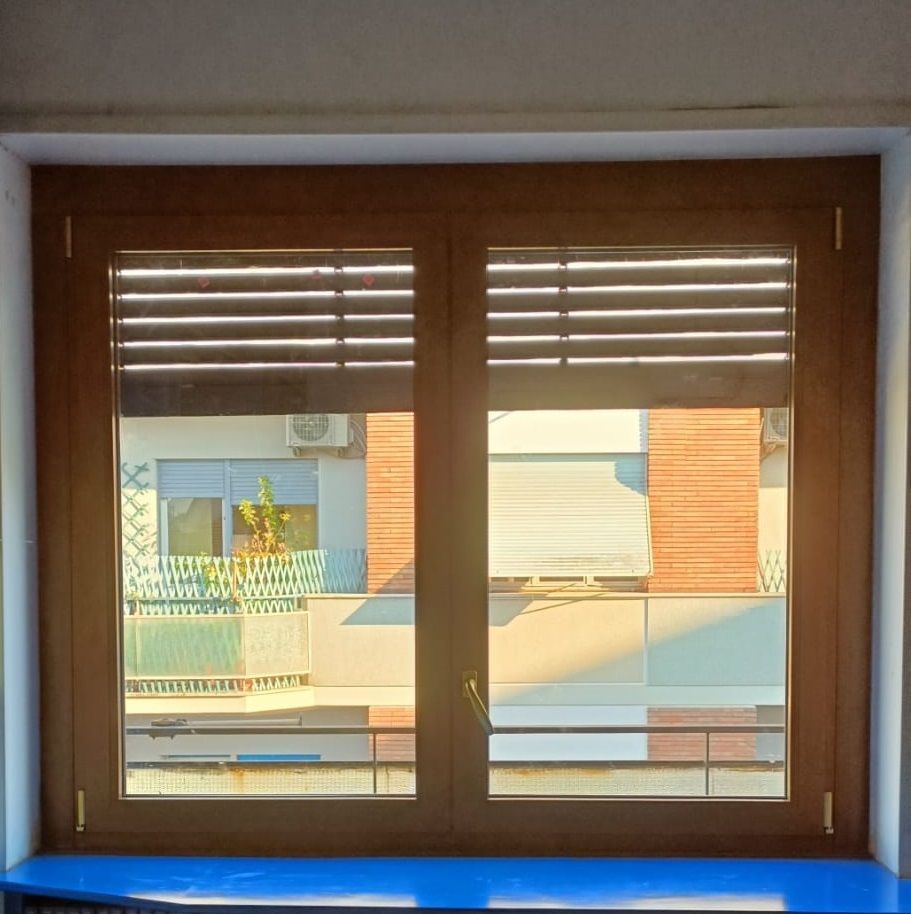 Finestra con telaio in legno e persiane orizzontali chiuse. Attraverso il vetro si vedono l'edificio e il cielo soleggiato.