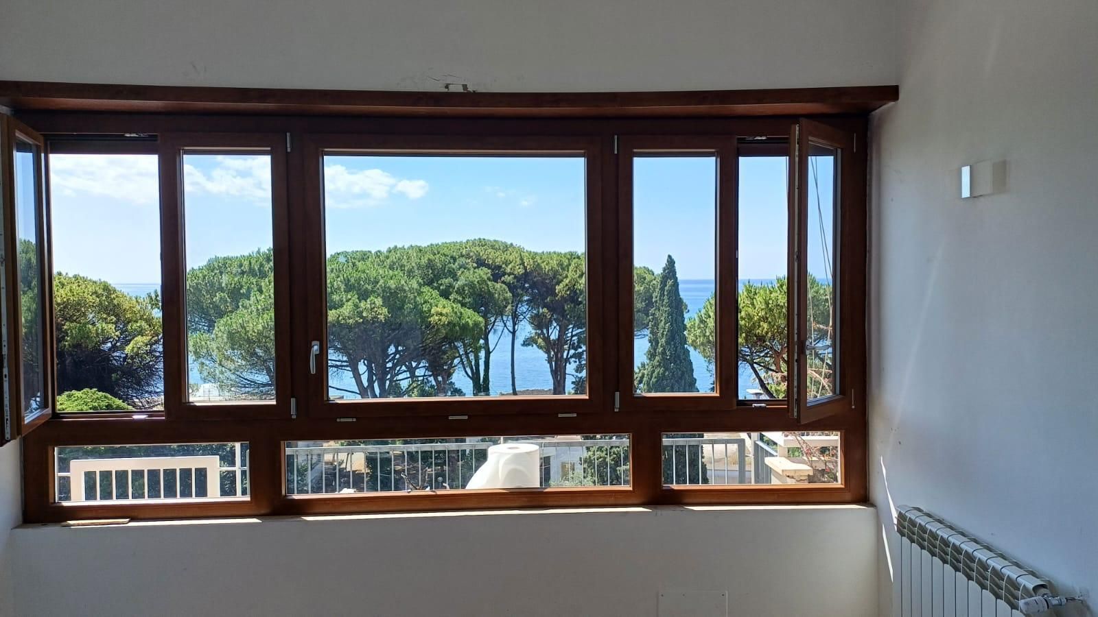 Finestre con cornice in legno con vista sull'oceano e rigogliosi alberi verdi sotto un cielo azzurro.