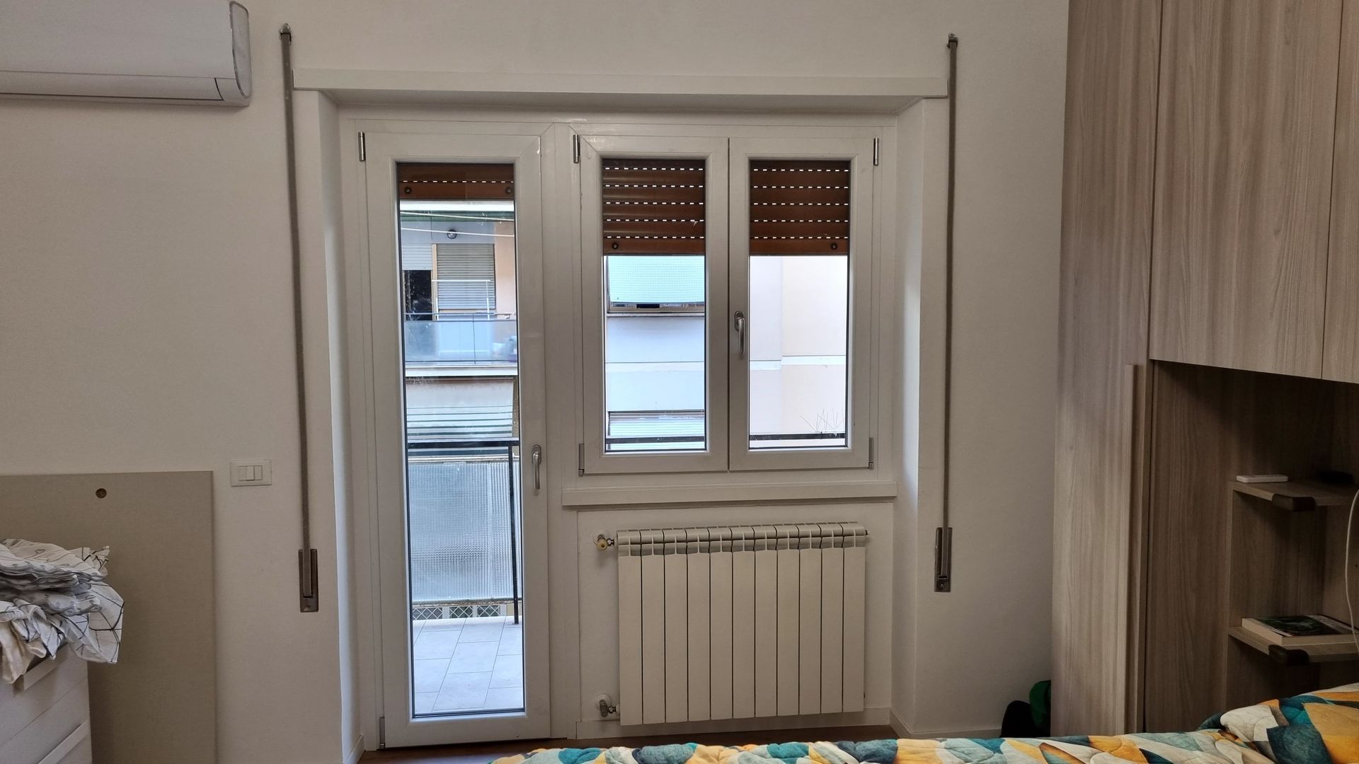 Una finestra e una porta bianche si aprono su un balcone, con un termosifone sotto la finestra, il tutto in una stanza dalle pareti bianche.
