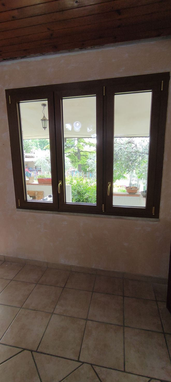 Una finestra marrone a tre vetri che si affaccia su un verde paesaggio esterno. La stanza ha pavimenti piastrellati e pareti color crema.