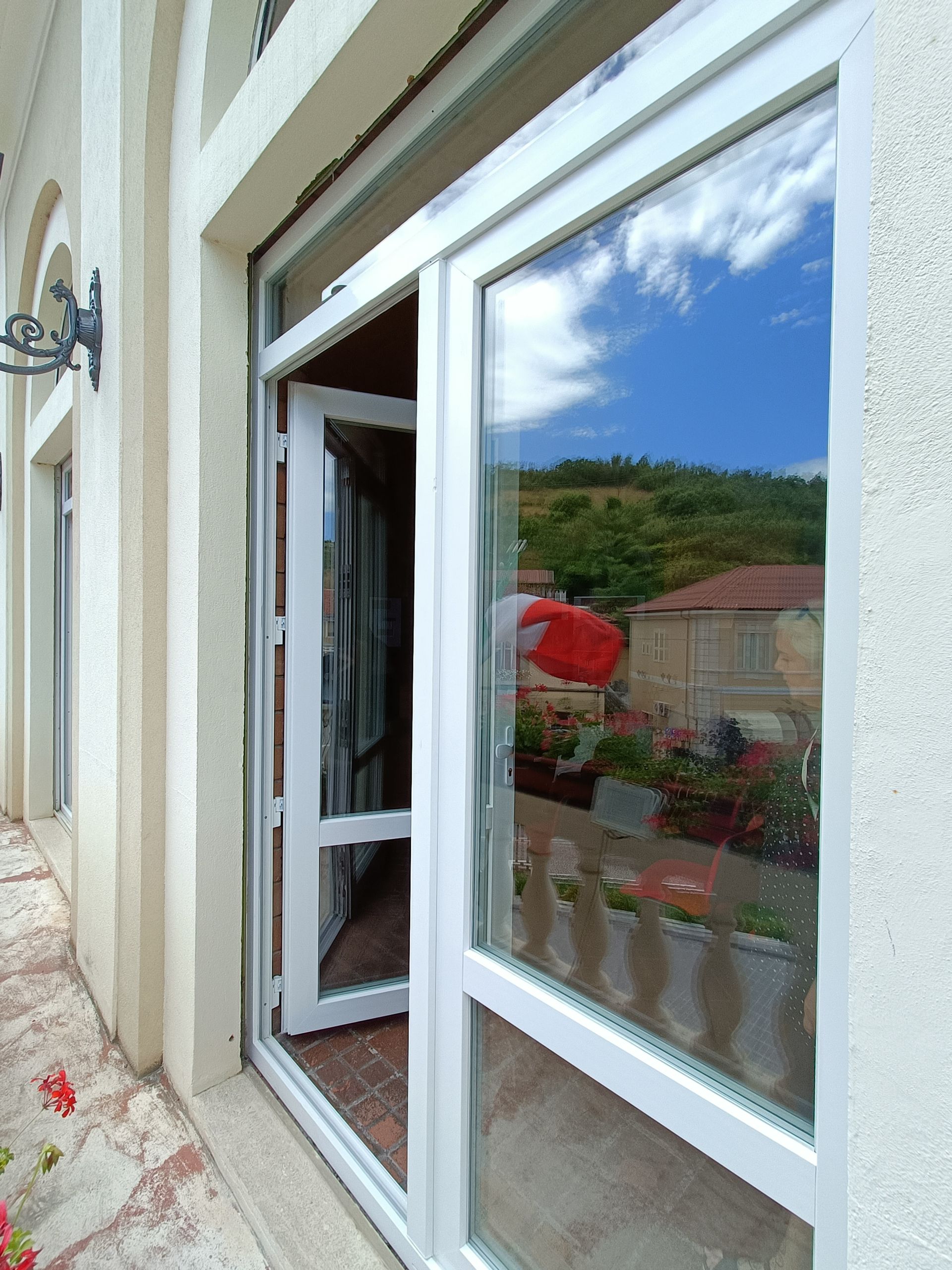 Porta a vetri con cornice bianca che si apre su un balcone. Riflette il cielo, la collina verde e l'edificio con i fiori rossi.