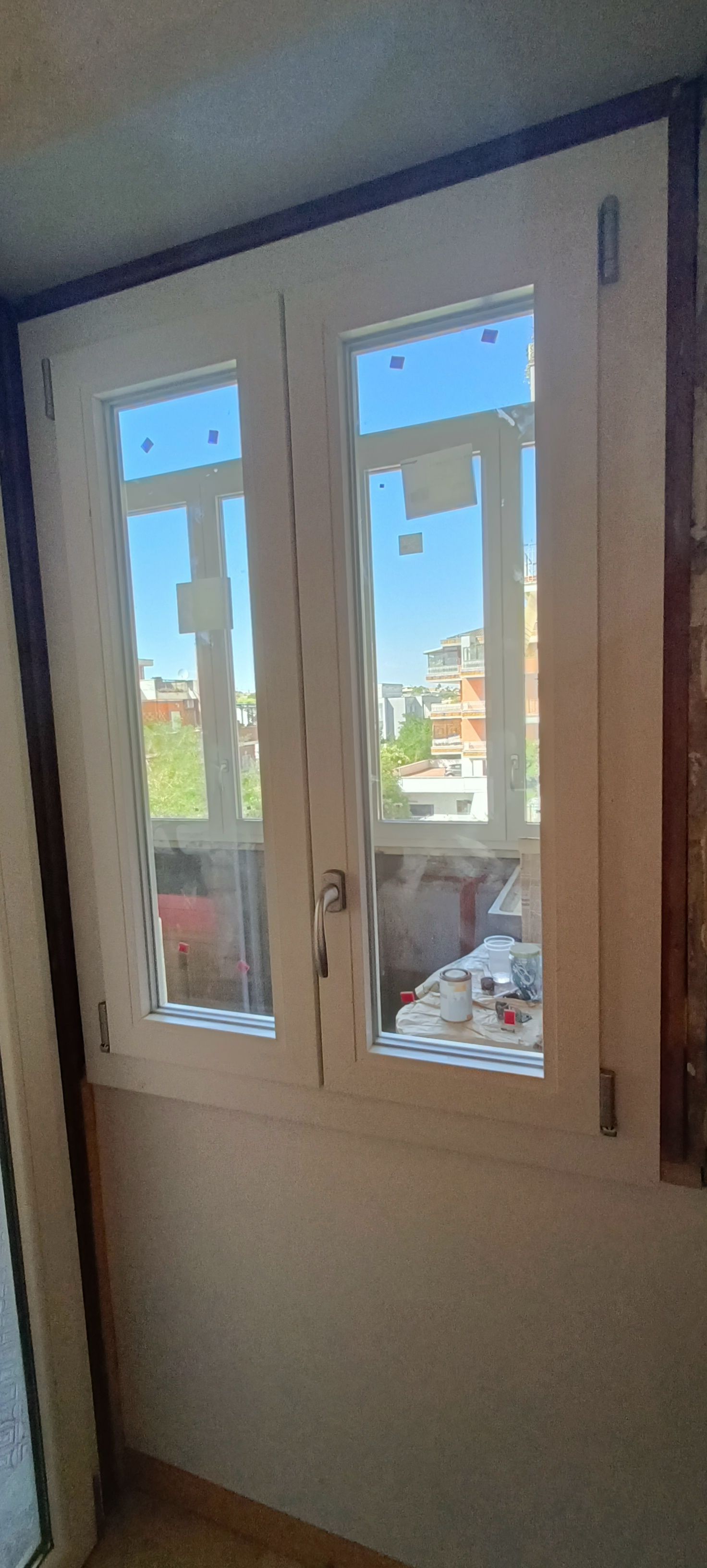 Finestra a due vetri, cornice bianca, aperta sul paesaggio urbano. Cielo azzurro e limpido visibile.