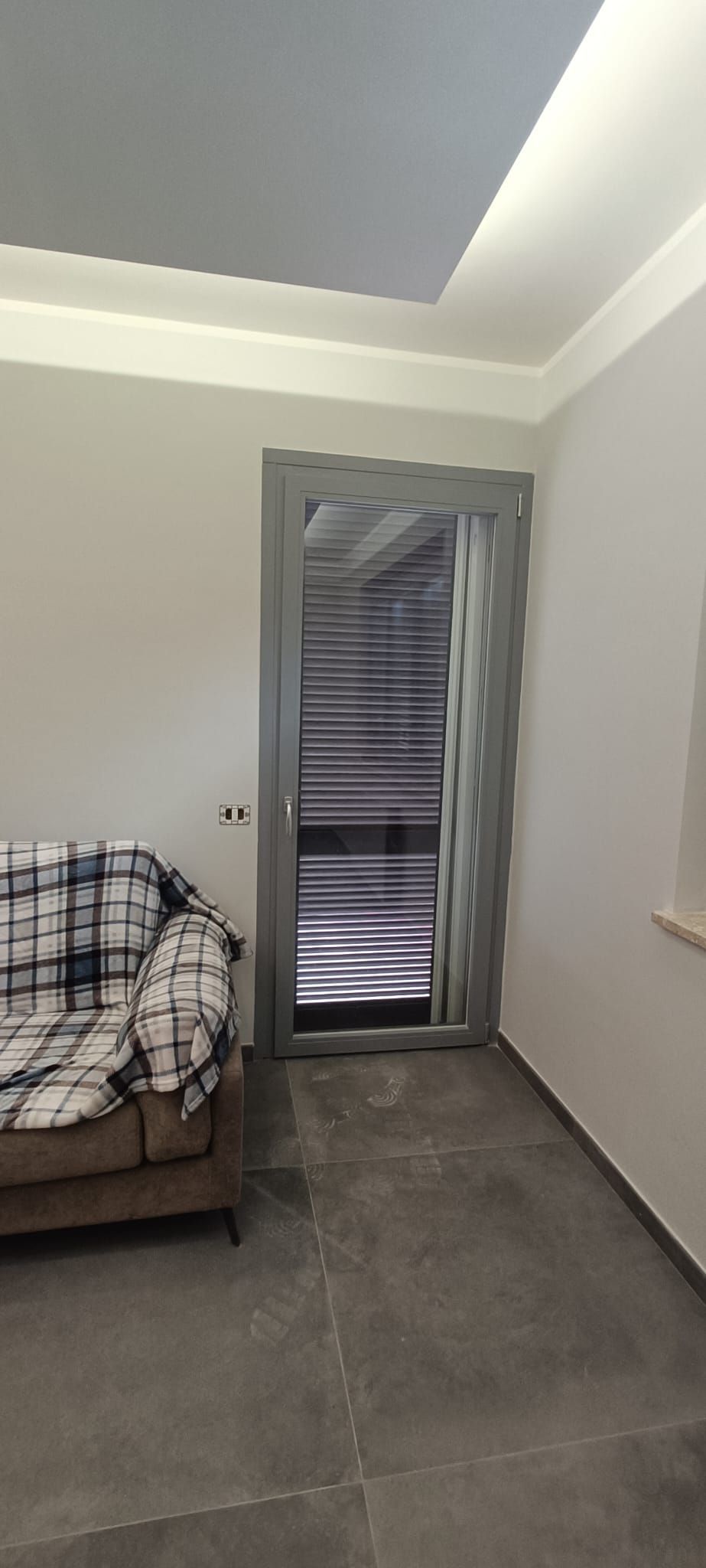 Stanza con divano, porta grigia con listelli e pavimento grigio. Il soffitto è grigio chiaro con finiture bianche.