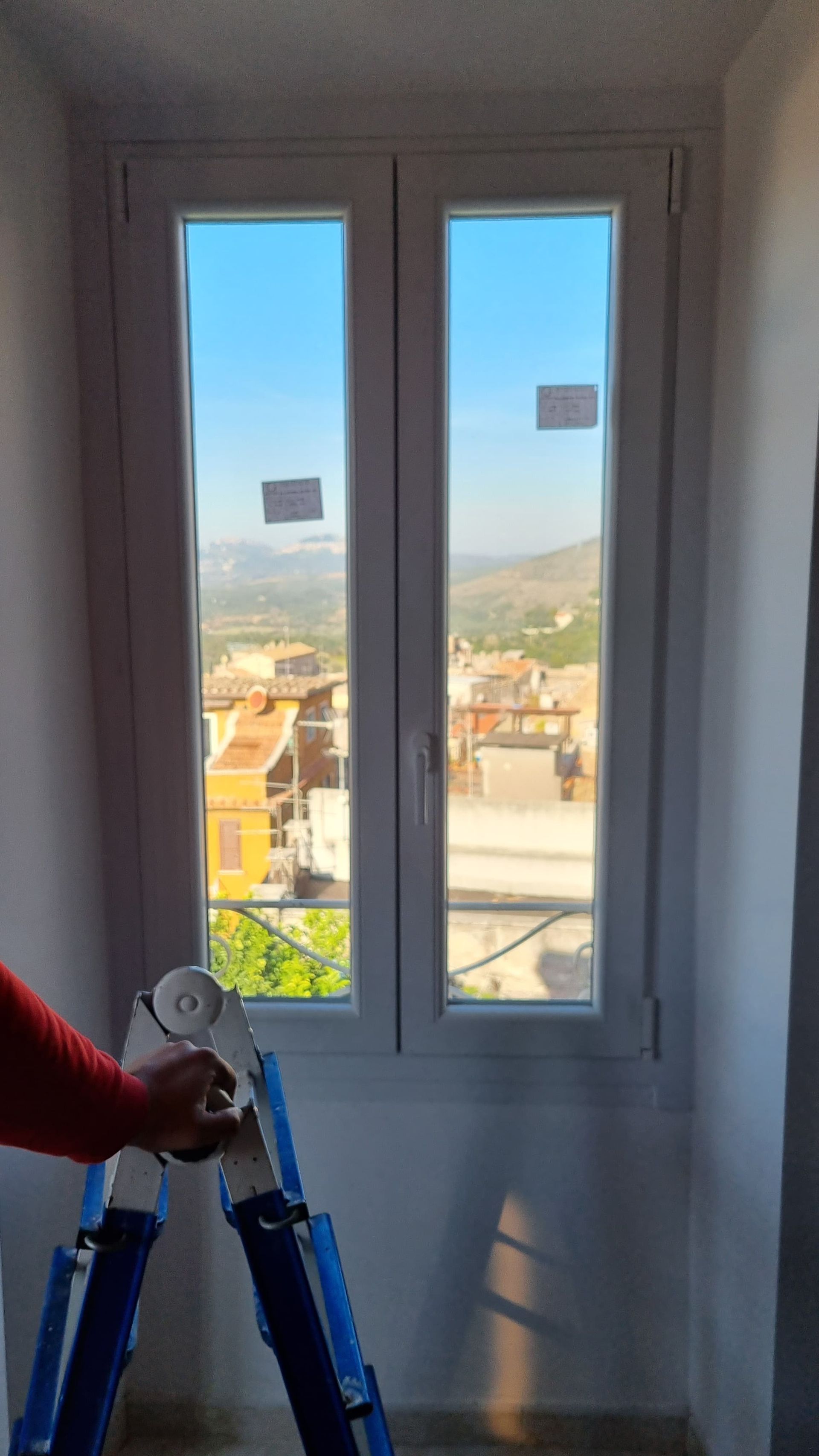 Una persona in piedi su una scala con in mano un attrezzo, guarda fuori dalla finestra verso una città in una giornata di sole.