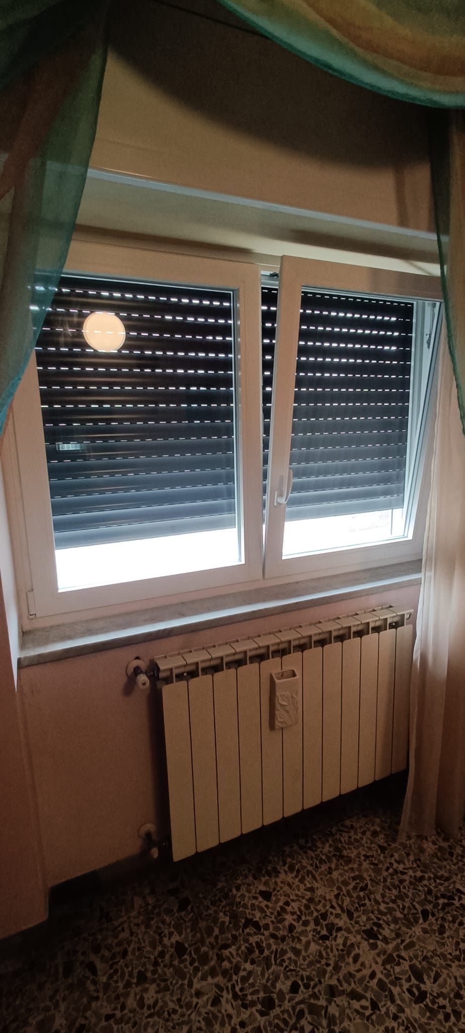 Finestra con persiane e termosifone sottostante. Sulla destra si vede una tenda.