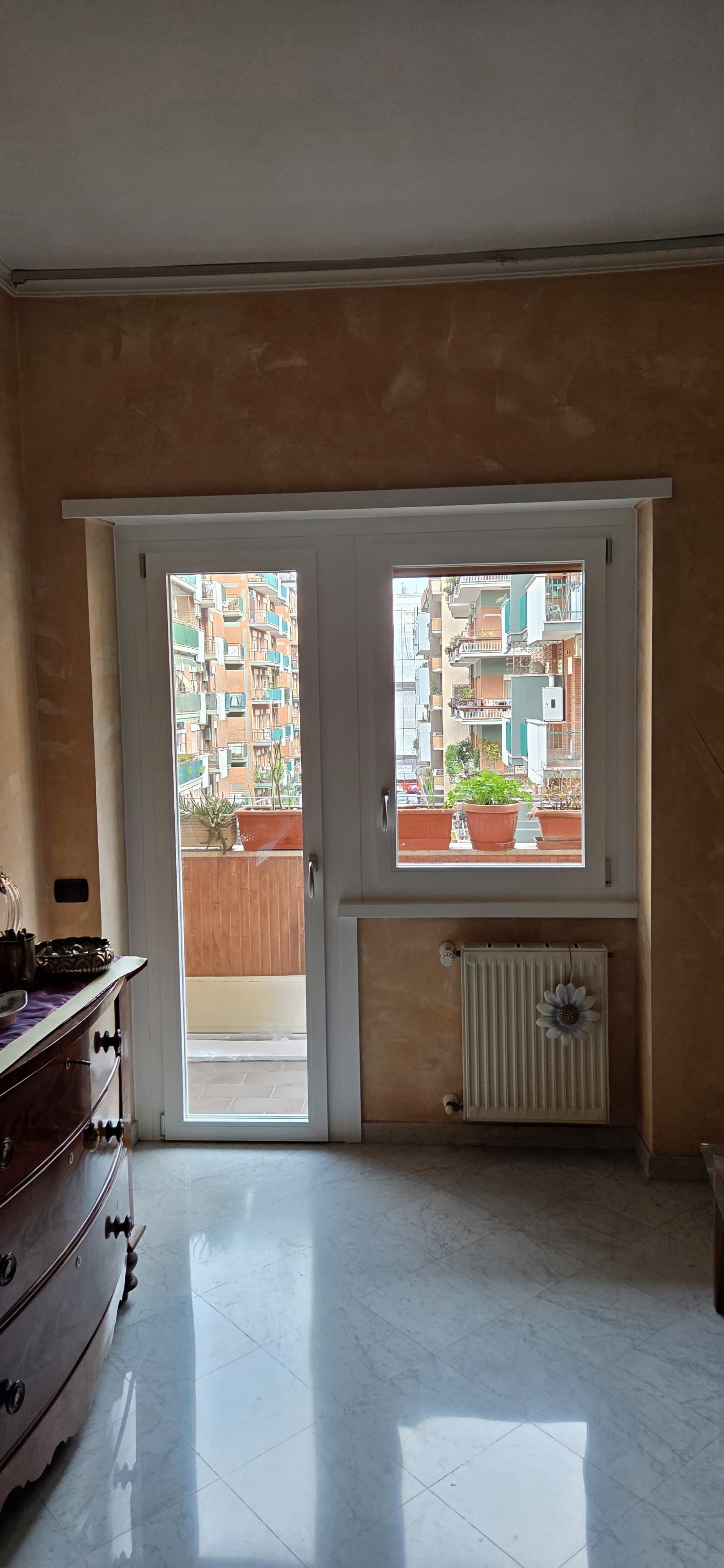 Stanza interna con porta che dà su un balcone con vista sulla città; mobile in primo piano, termosifone sotto la finestra.