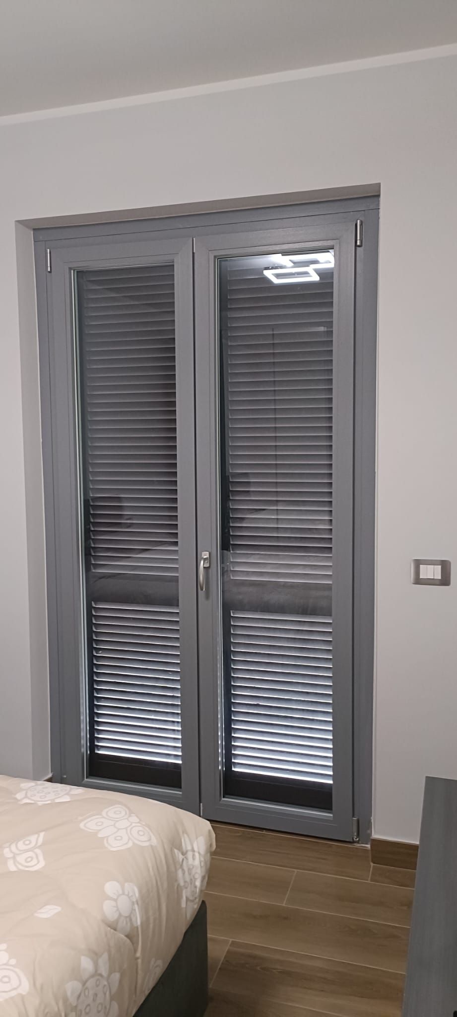 Persiane grigie visibili attraverso le porte a vetri. Si vedono un letto e una presa a muro.