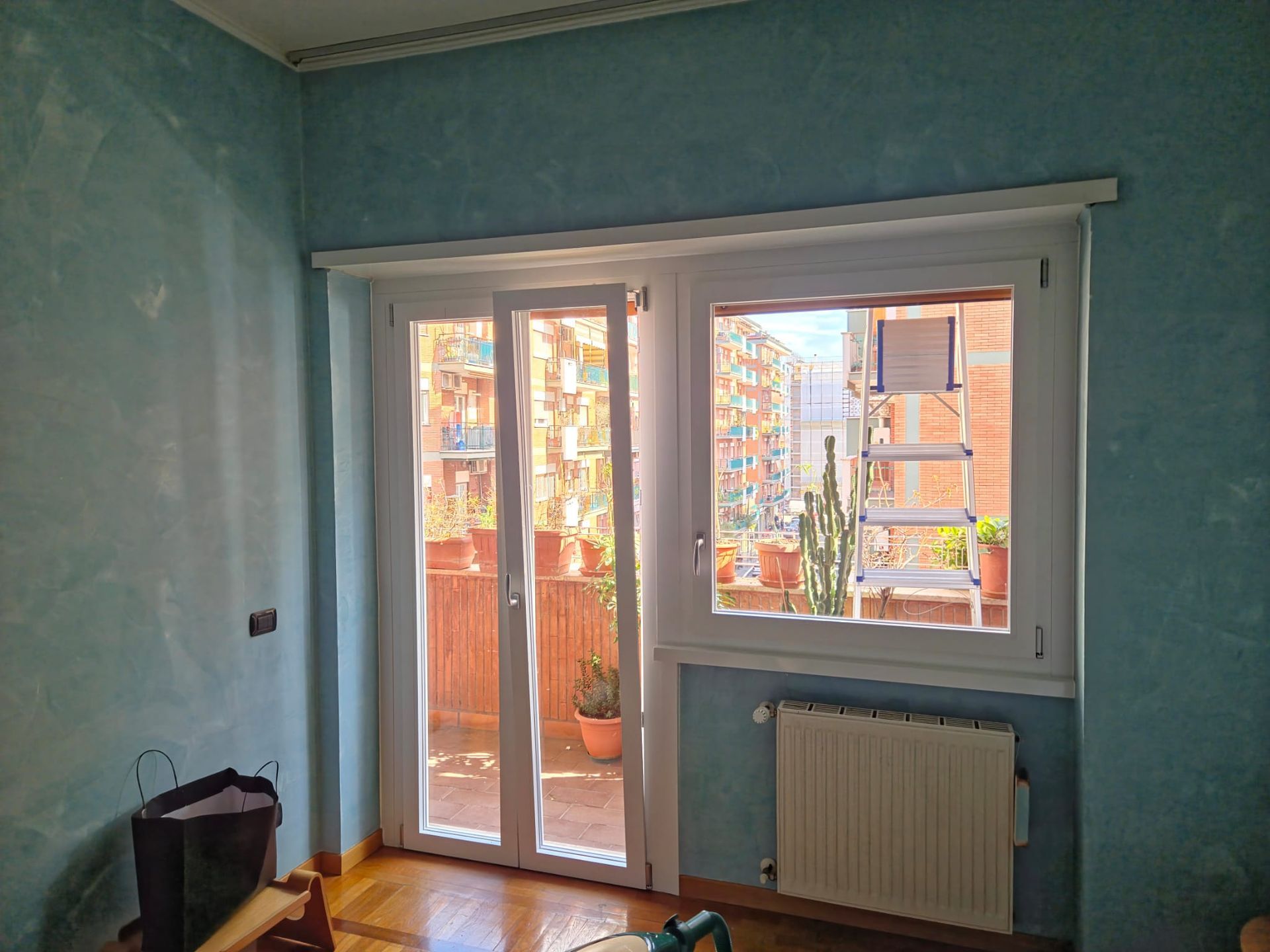 Finestra e porta con cornici bianche, con vista su un balcone e sul paesaggio urbano. Pareti blu, termosifone bianco e pavimenti in legno.