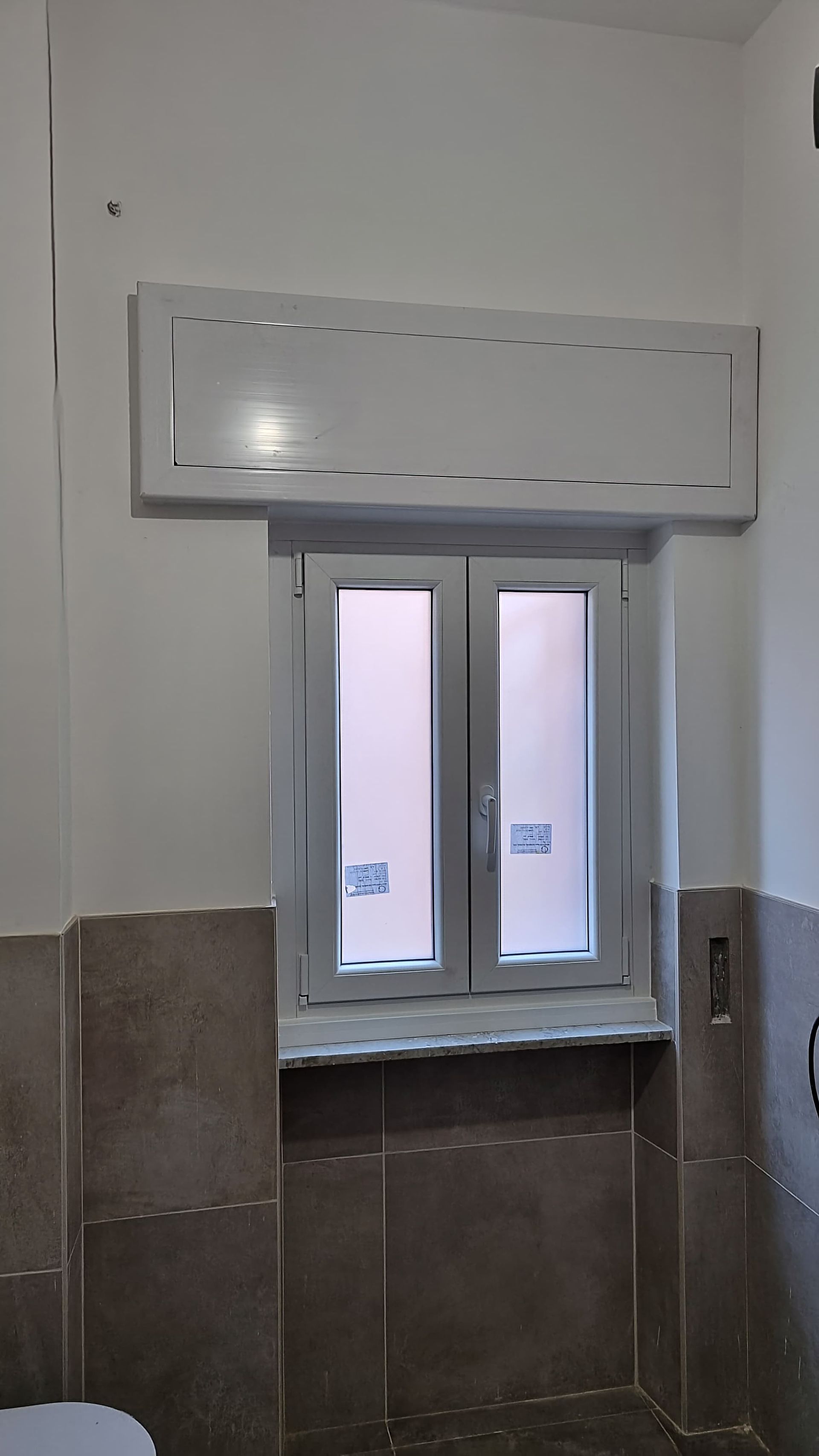 Finestra con cornice bianca in un bagno con piastrelle grigie e un mobile contenitore bianco sopra la finestra.