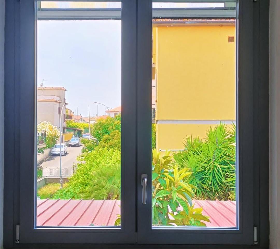 Finestra grigia che si affaccia su una strada con auto, alberi e un edificio giallo. Giornata luminosa e soleggiata.