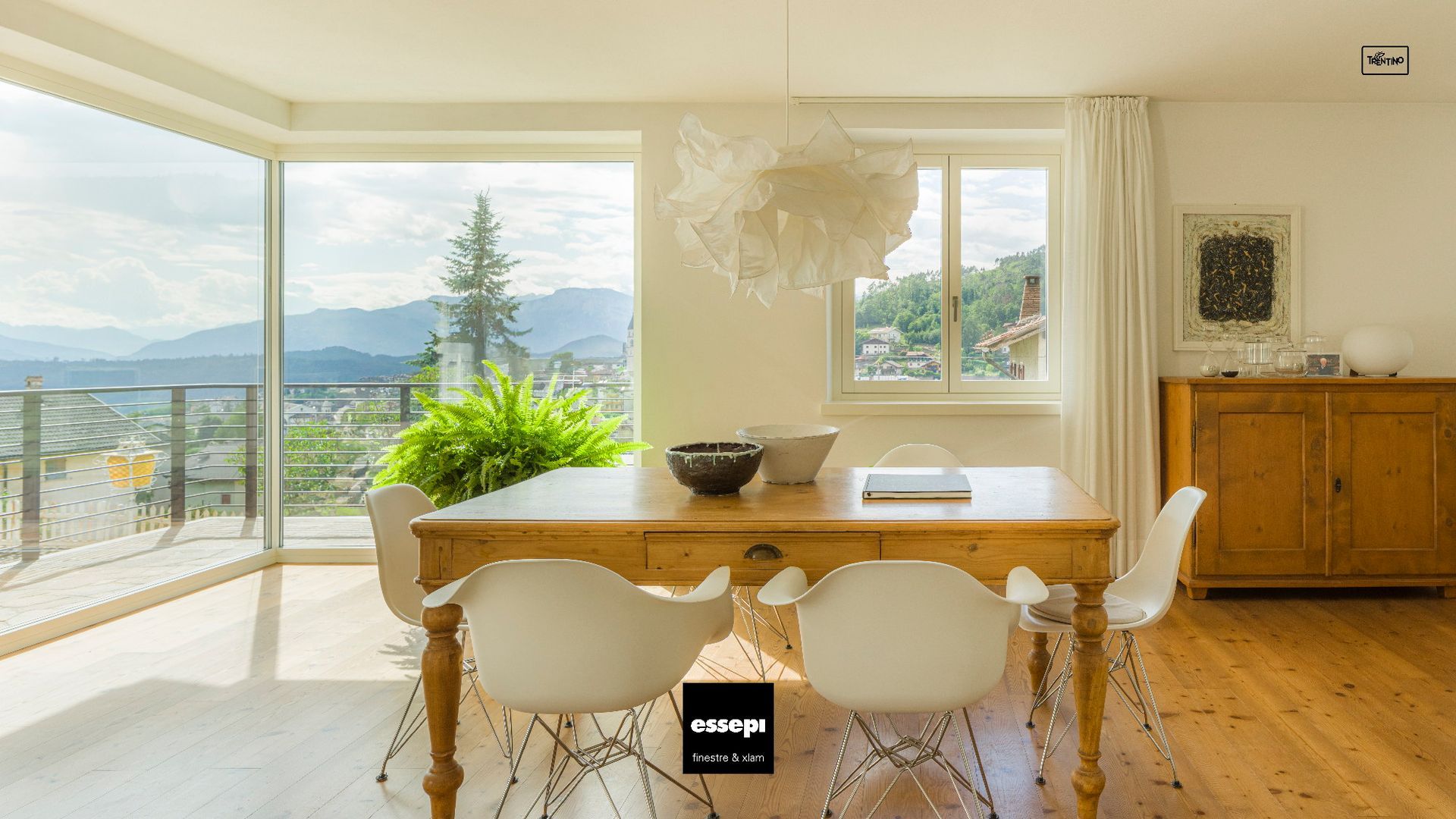 Sala da pranzo con tavolo, sedie bianche, ampie finestre con vista sulle montagne e lampadario decorativo.