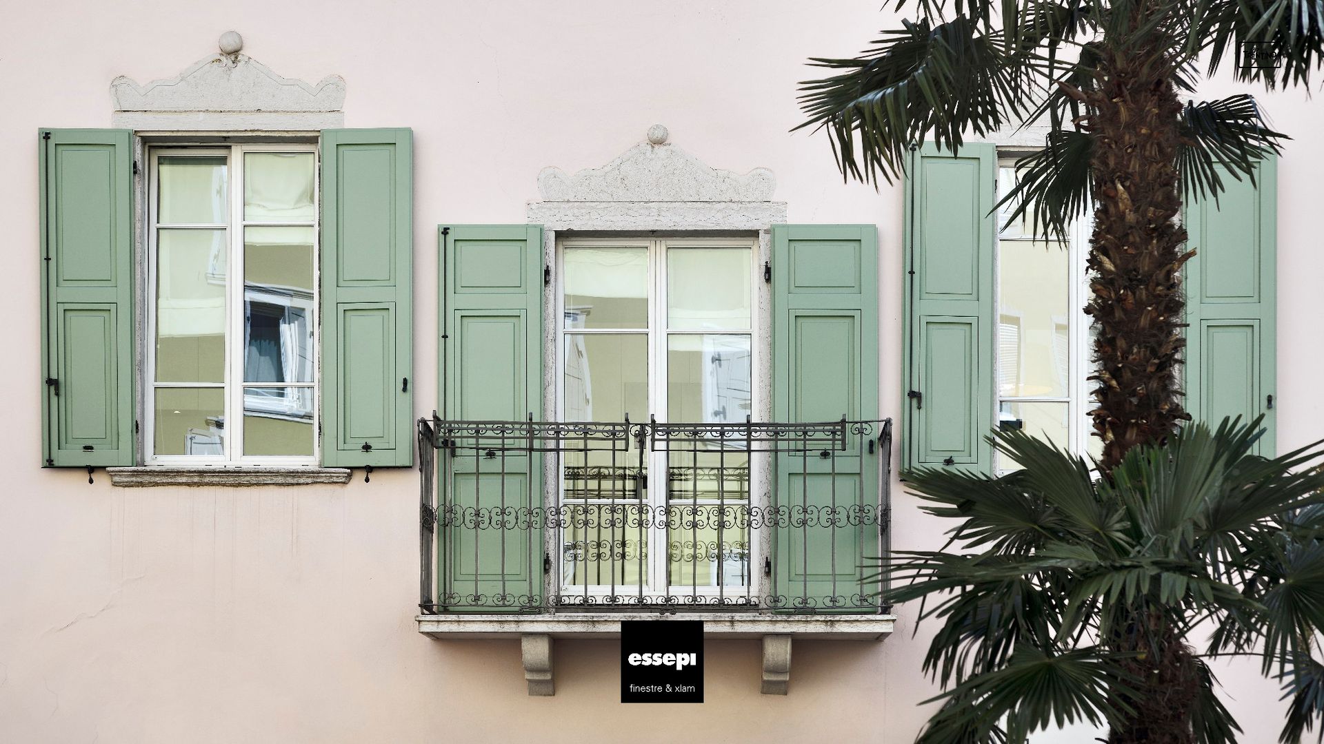 Edificio rosa con tre finestre, persiane verde chiaro e un balcone con ringhiera in ferro nero, palma.
