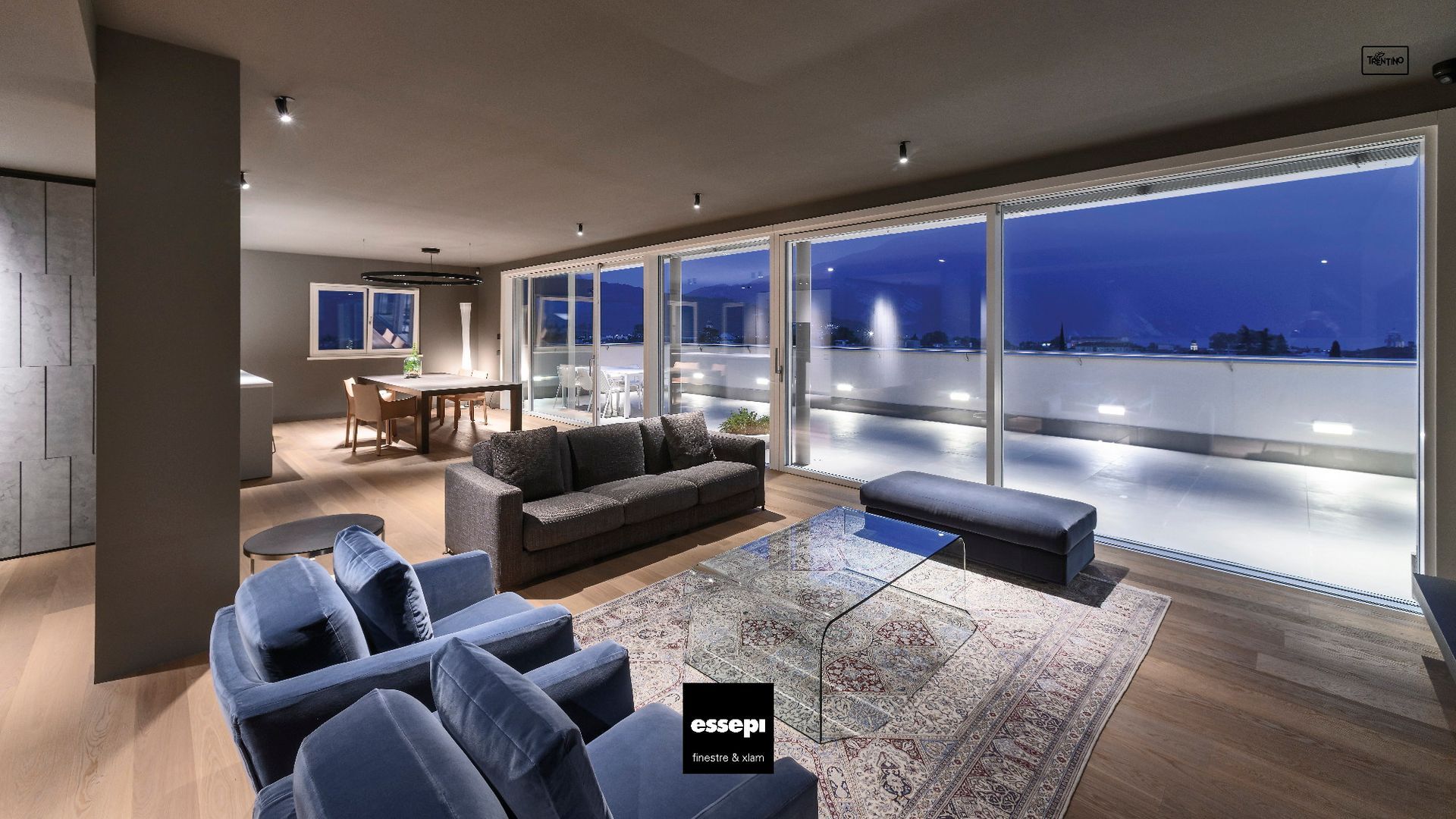 Moderno soggiorno con ampie finestre che si affacciano sulla città al tramonto. Presenta sedute blu, tappeto e pavimento in legno. Infissi in legno