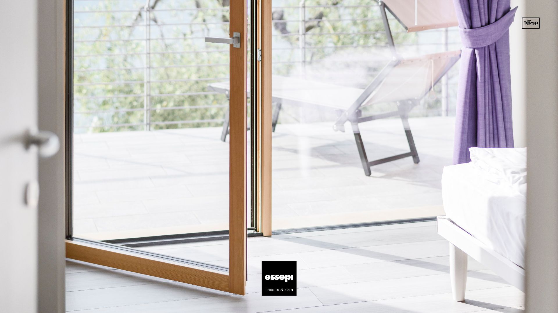 Porta scorrevole in vetro che si apre su un balcone, tenda lilla, telaio in legno, luce solare intensa.