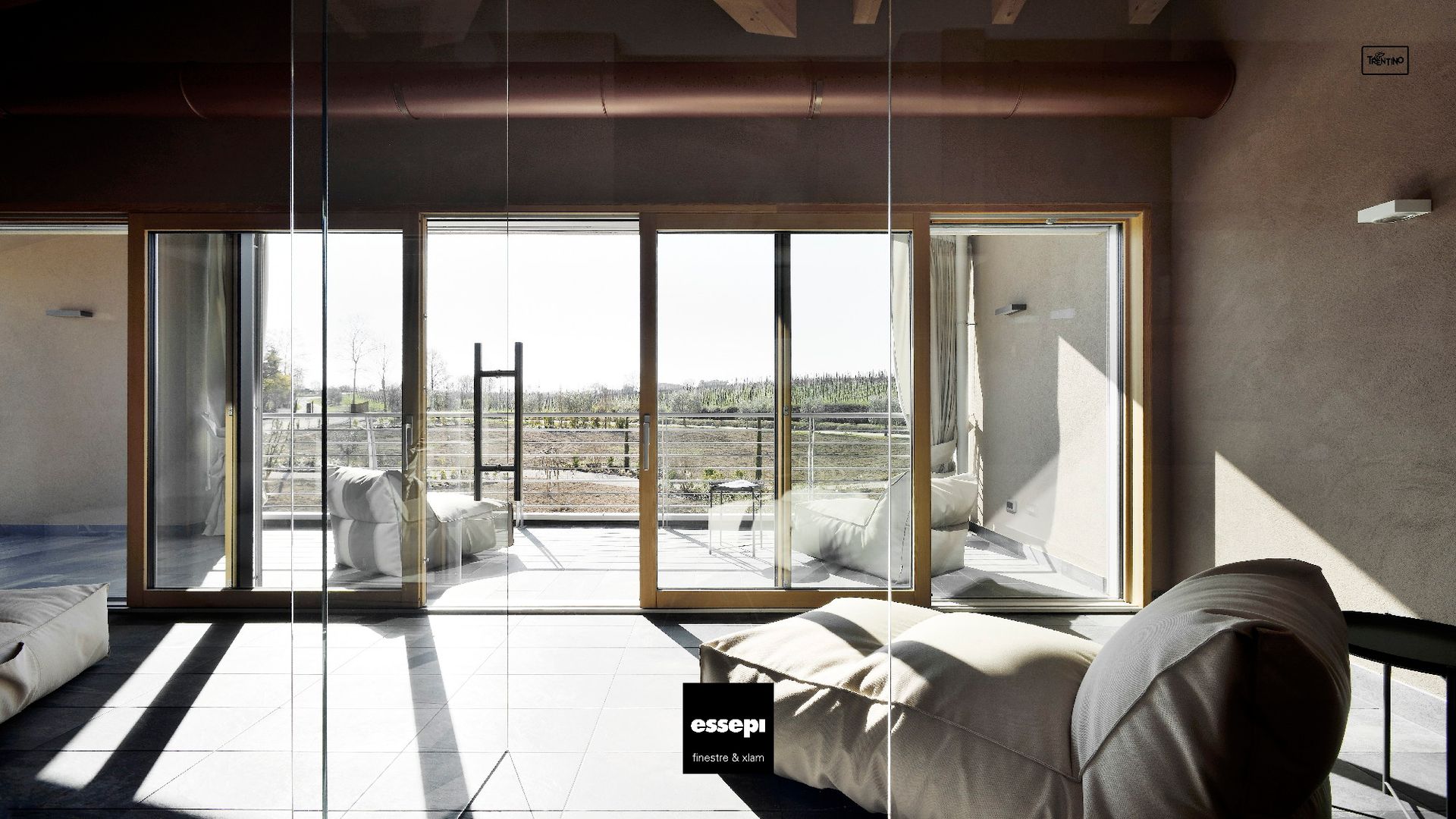 Camera con ampie finestre che si affacciano sul paesaggio. Infissi in legno, pouf bianchi, luce solare.