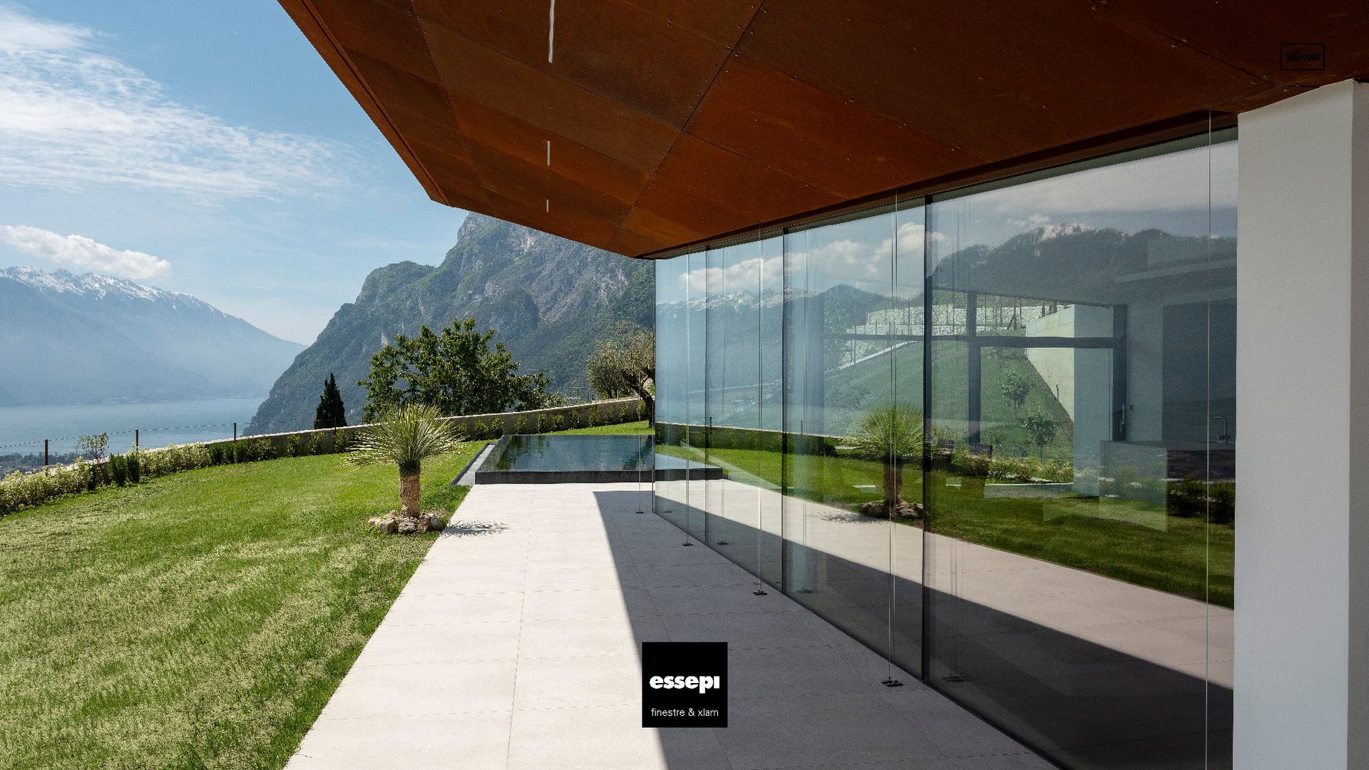 Moderno edificio con pareti in vetro, vista sul lago e sulle montagne, caratterizzato da un tetto marrone e da un'area erbosa.
