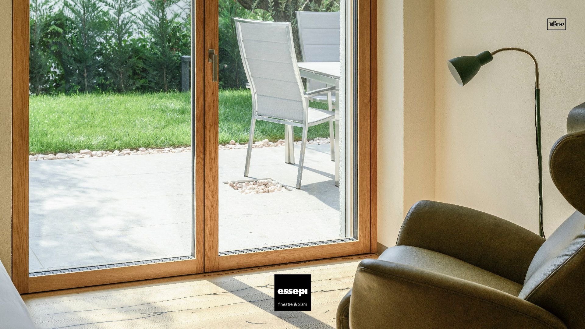 Le porte-finestre con cornice in legno si aprono su un patio con tavolo e sedie bianche. All'interno, una sedia e una lampada.