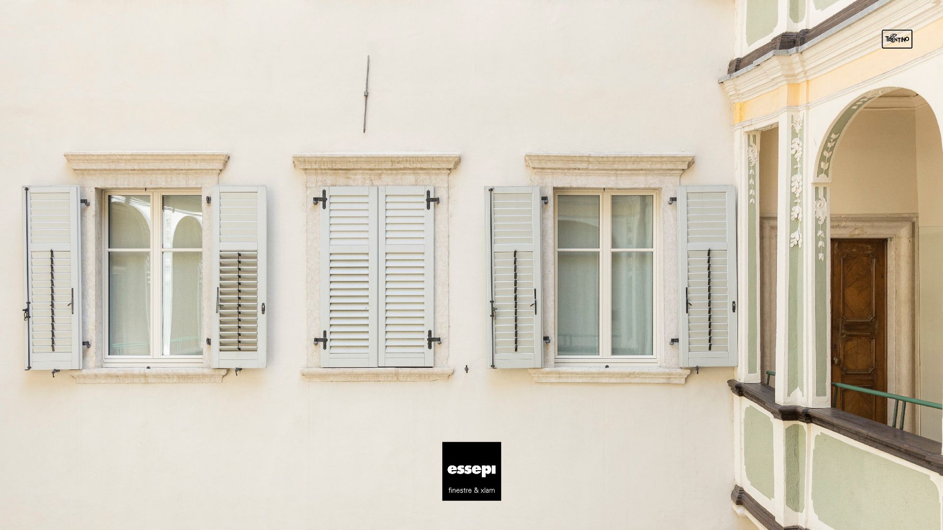 Tre finestre bianche con persiane aperte su una parete color crema; sulla destra è parzialmente visibile un portale ad arco decorativo.