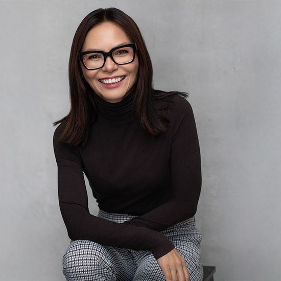 Woman with dark hair, glasses, black turtleneck, and patterned pants smiles against a gray background.