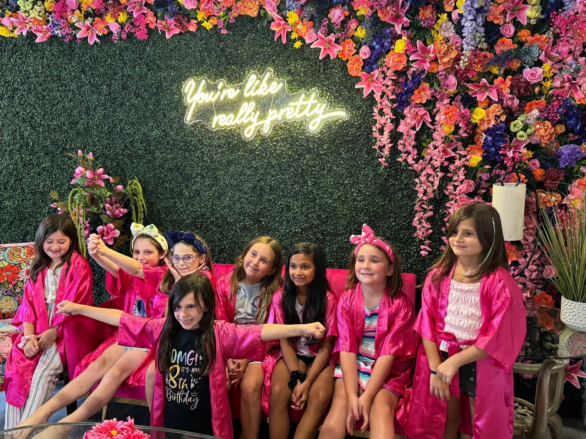 A group of young girls are sitting on a couch in front of a flower wall.