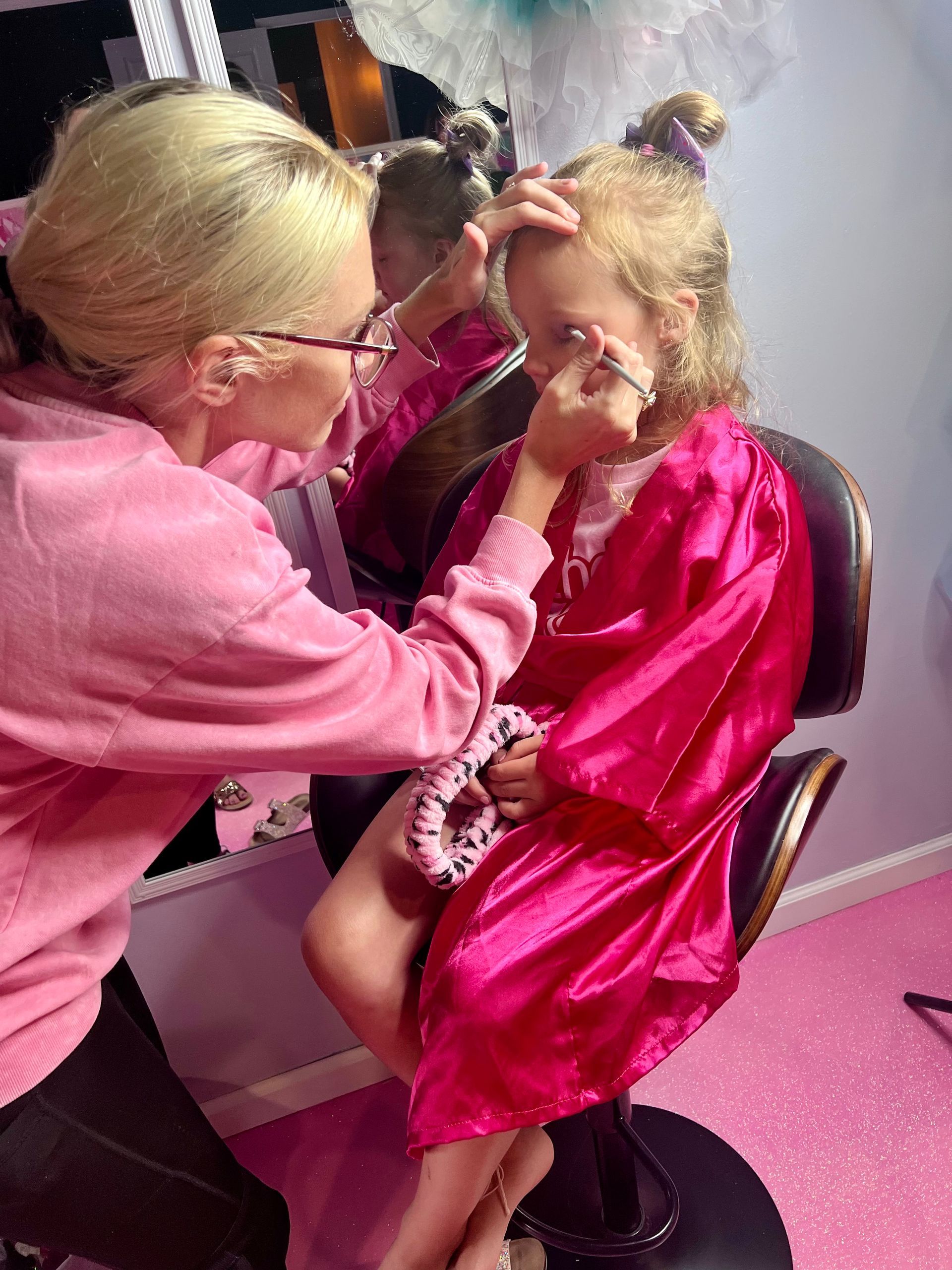A woman is applying makeup to a little girl in a pink robe.