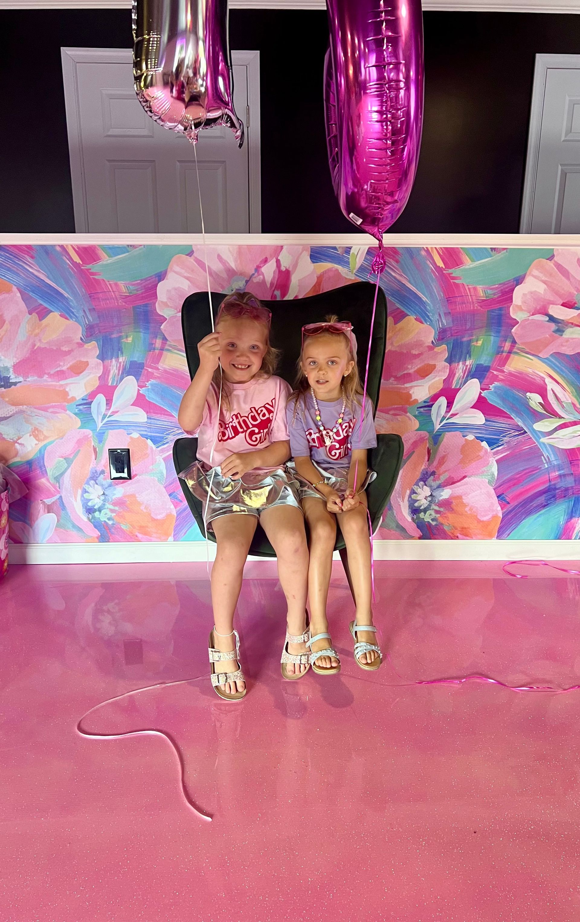 Two little girls are sitting on a chair in front of a pink wall with balloons.