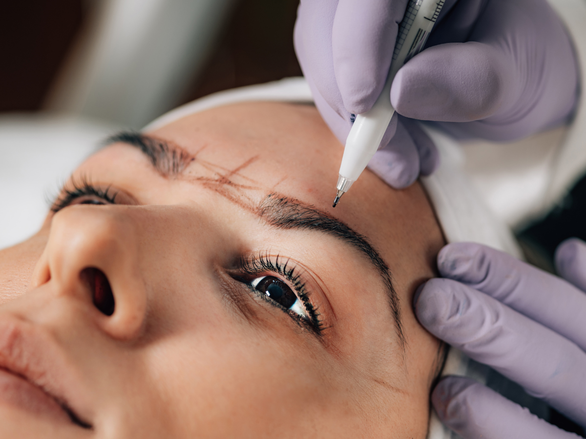 A person getting their eyebrows measured and outlined for a cosmetic procedure. Close-up with gloved hands and a pen.