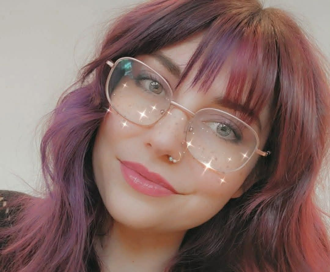 Close-up of a Sheylir with wavy burgundy hair, glasses, and a soft smile against a light background