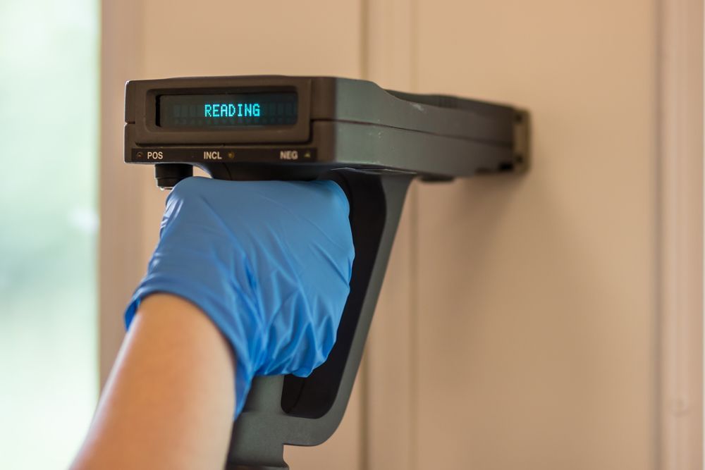 A person wearing blue gloves is holding a device on a wall.