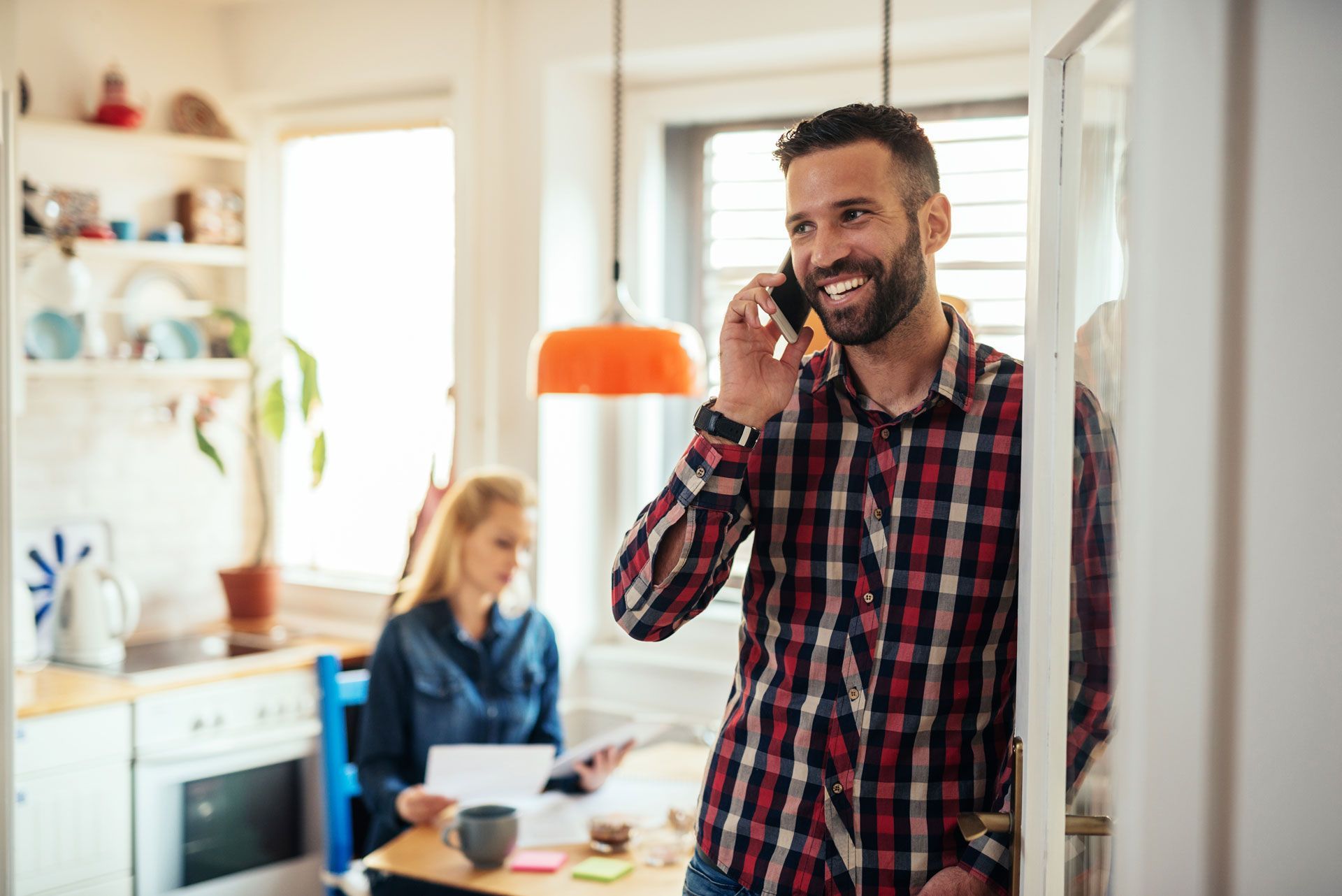 A man is talking on a cell phone in a kitchen while a woman sits at a table.