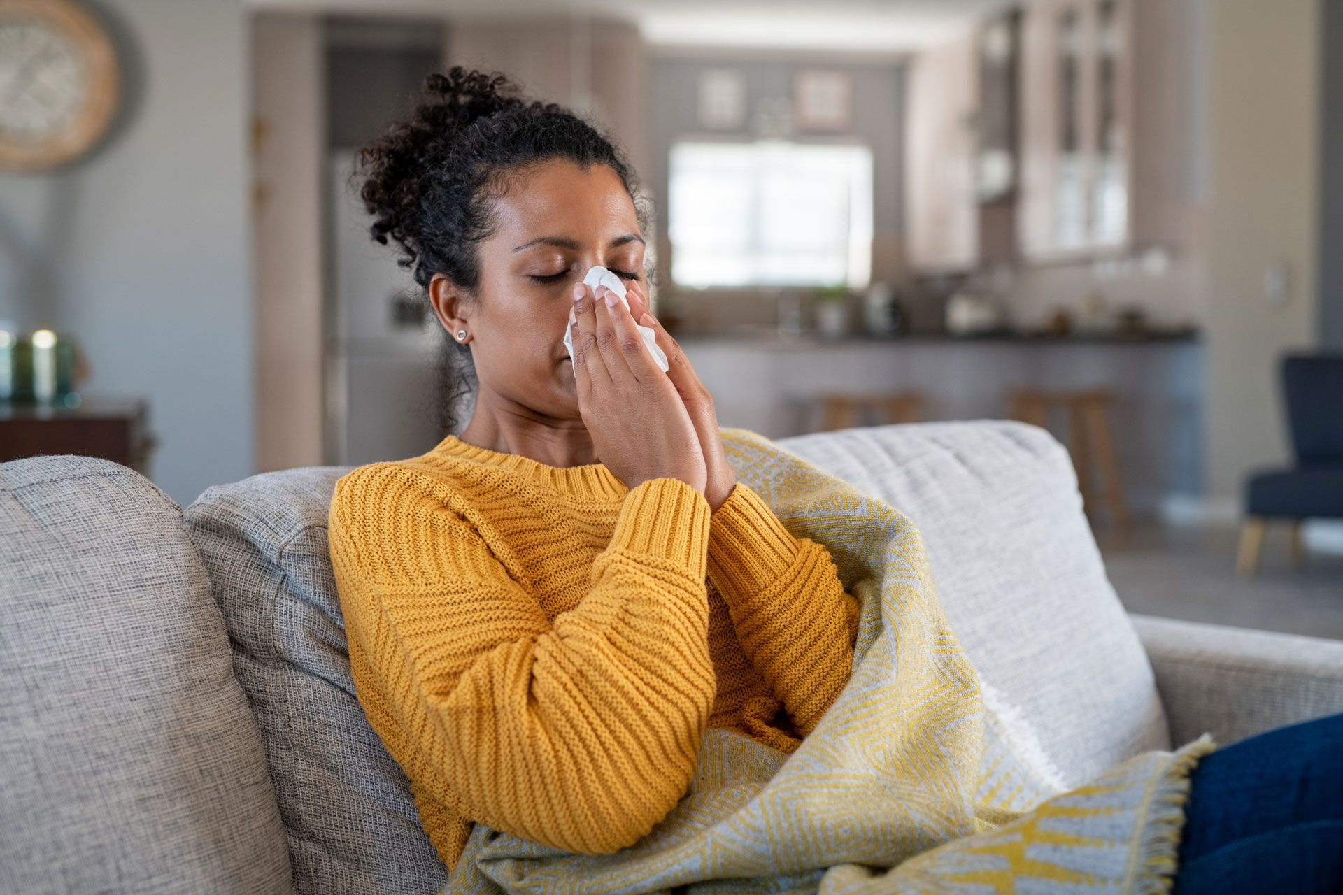 A woman is sitting on a couch blowing her nose into a napkin.
