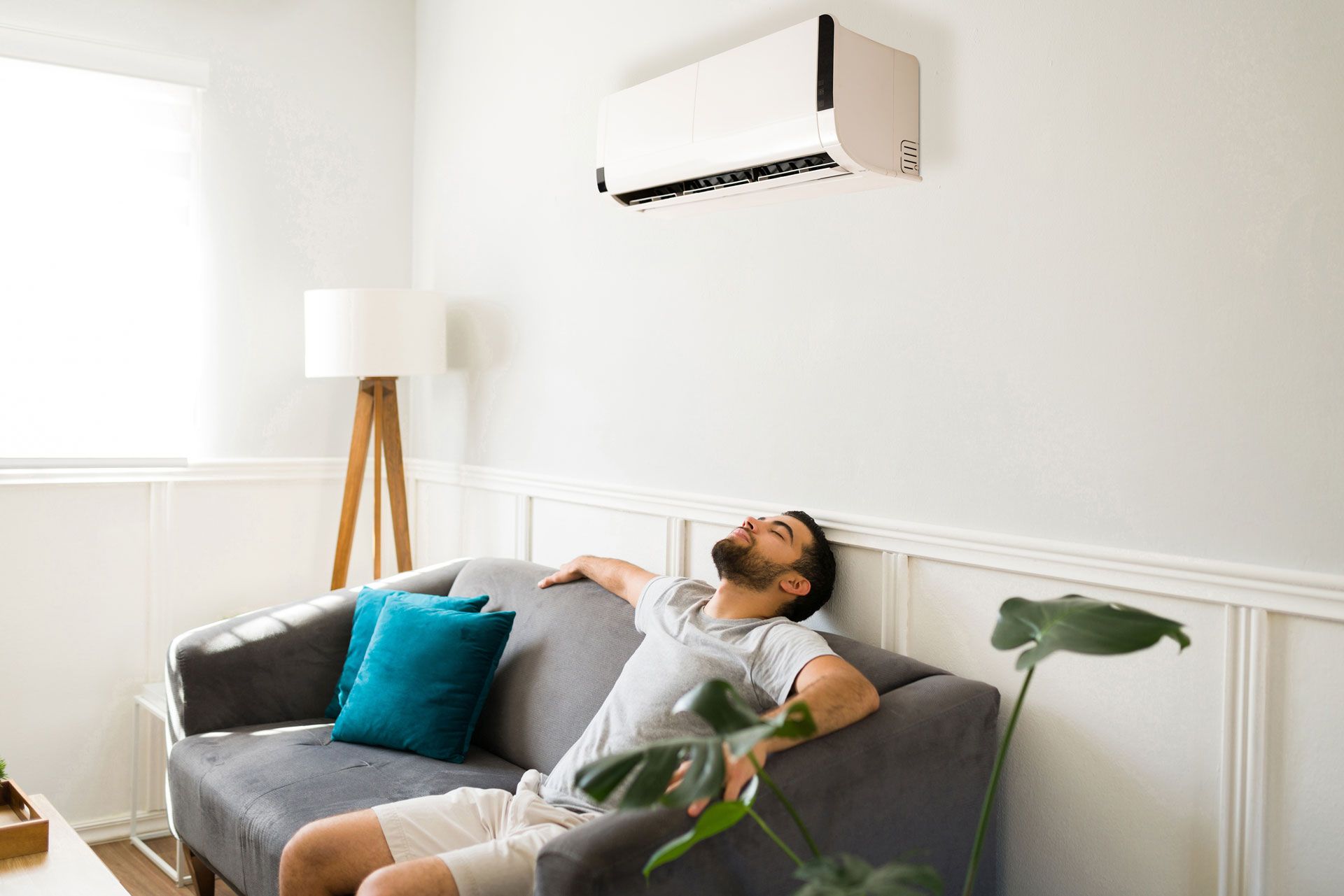 A man is sitting on a couch in a living room under an air conditioner.