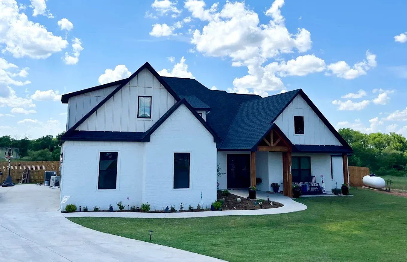 White farmhouse with black roof and trim on a sunny day. Green lawn and blue sky.