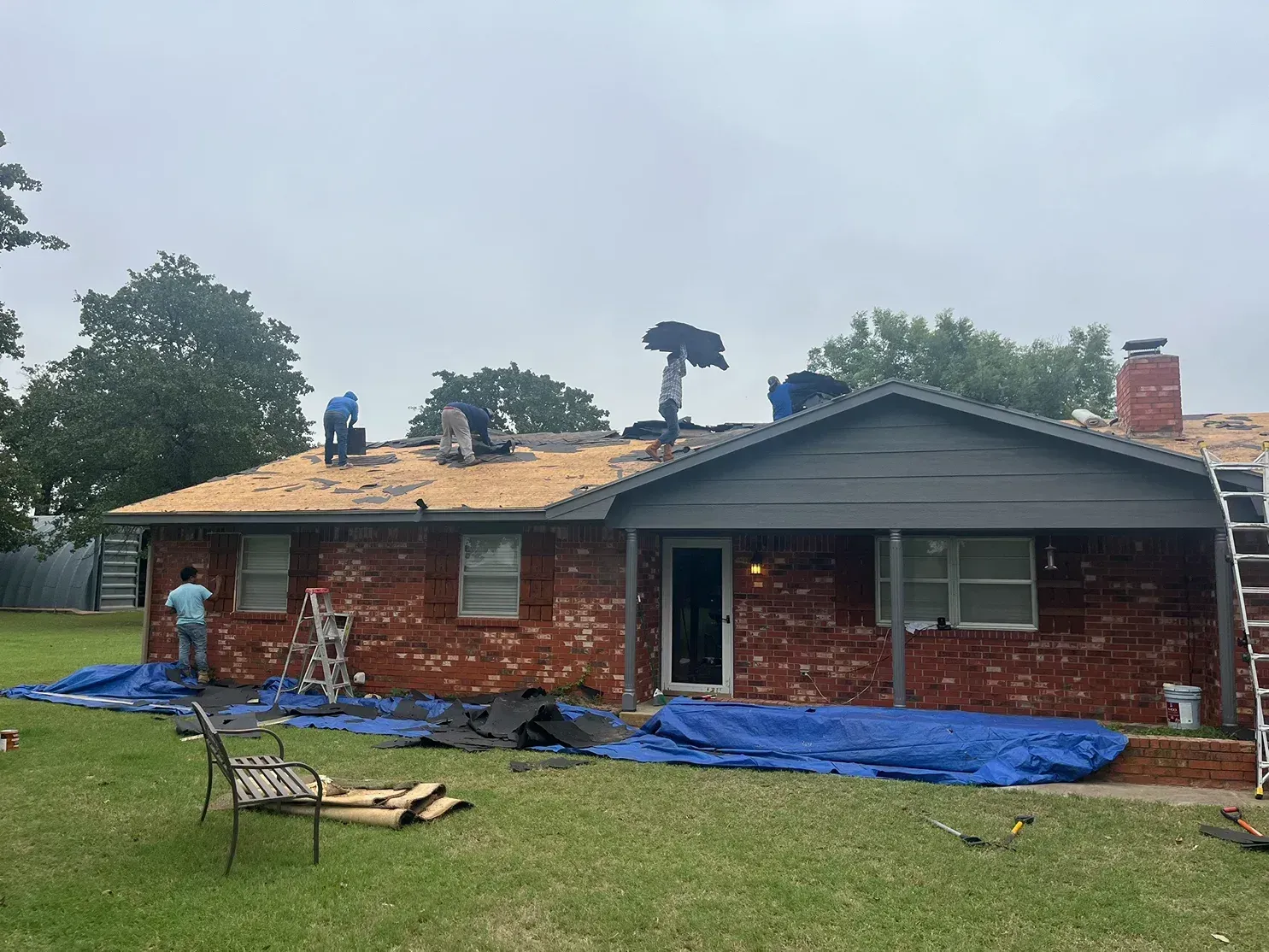 Roofers working on a red brick house under overcast skies, with ladders and tarps.