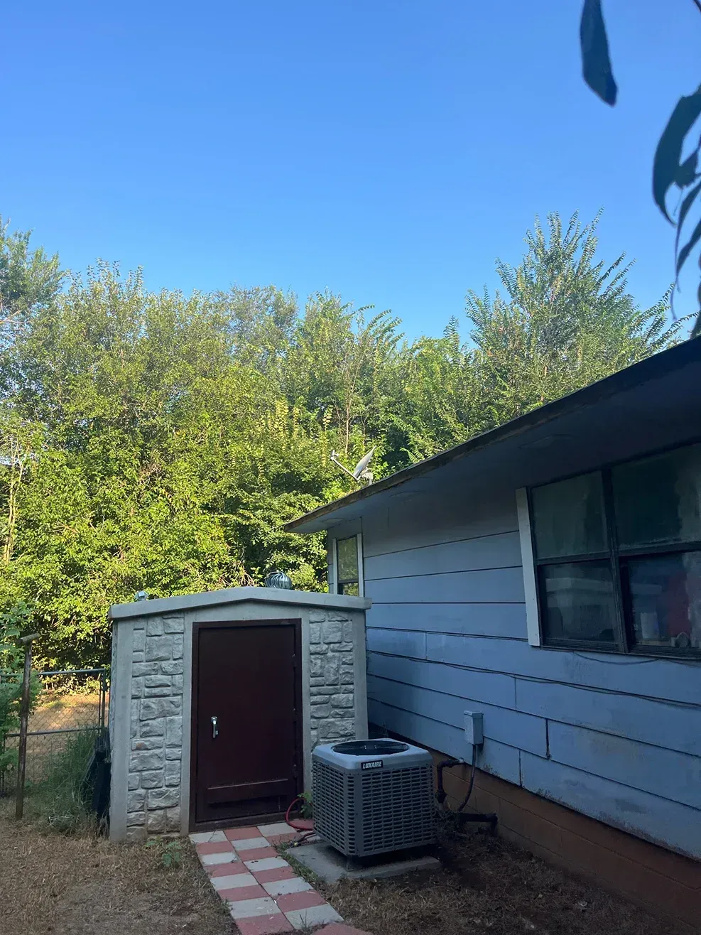 Small stone shed with a dark door, next to a light blue house, under a clear blue sky.
