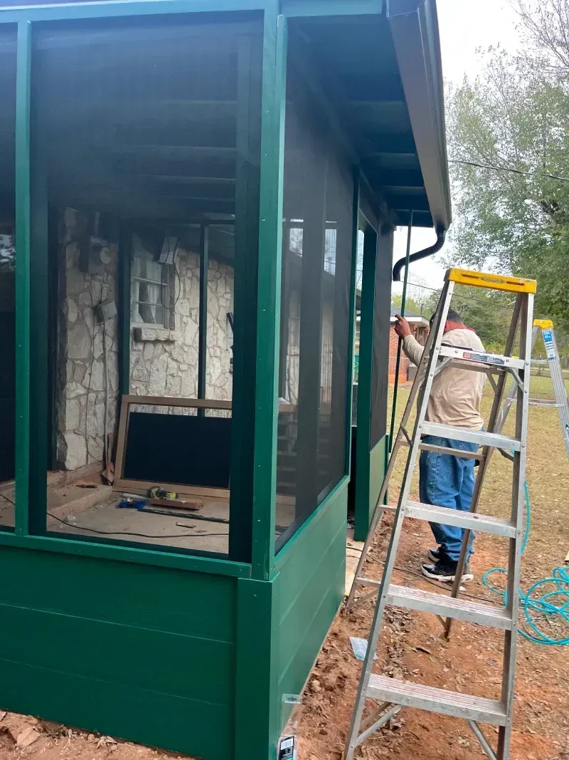 Green screened-in porch under construction; person on ladder, working.