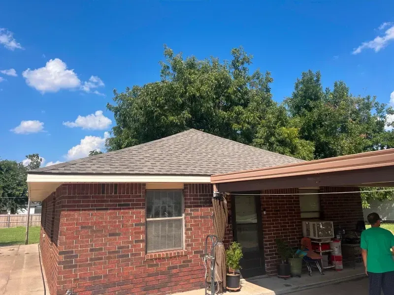 Brick house with a new gray shingle roof and carport. A man in green stands near the carport. Blue sky with clouds.