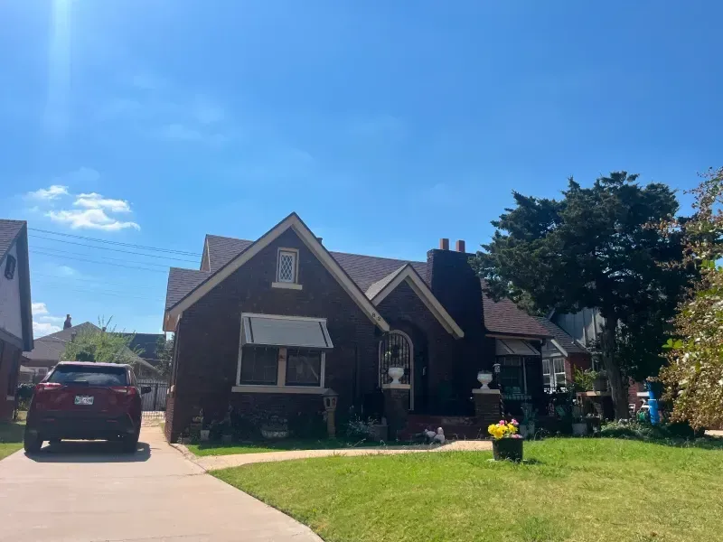 Brown brick house with a brown roof, white awning, and a small front yard with green grass under a blue sky.