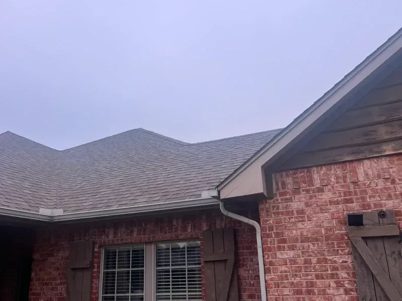 Brick house with brown roof, shutters, and gutters under a cloudy sky.