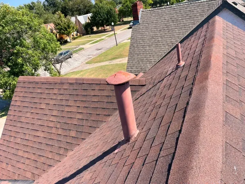 Brown shingled roofs with a red vent pipe, viewed from above, with houses and trees in the background.