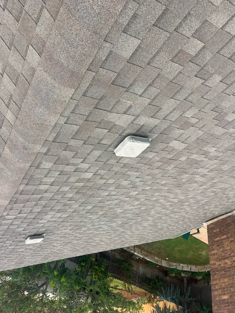 Asphalt shingle roof with two white square objects on the surface. Green foliage and building in the background.