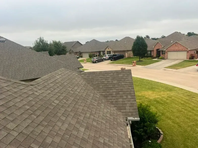 Roofs of suburban homes with brown shingles. Overcast sky. Some cars and lawns are visible.