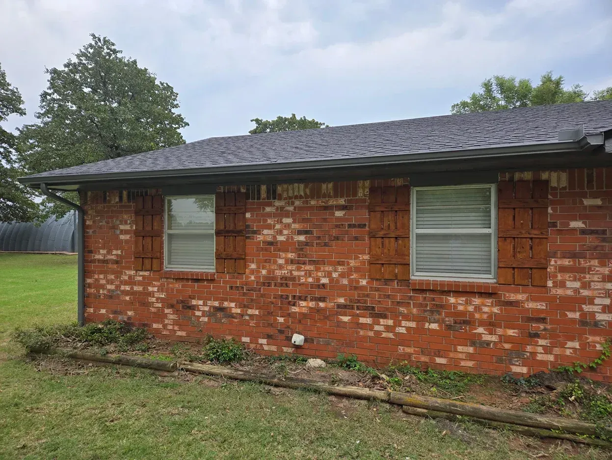 Red brick house with two windows, dark roof, and green grass. Cloudy sky.